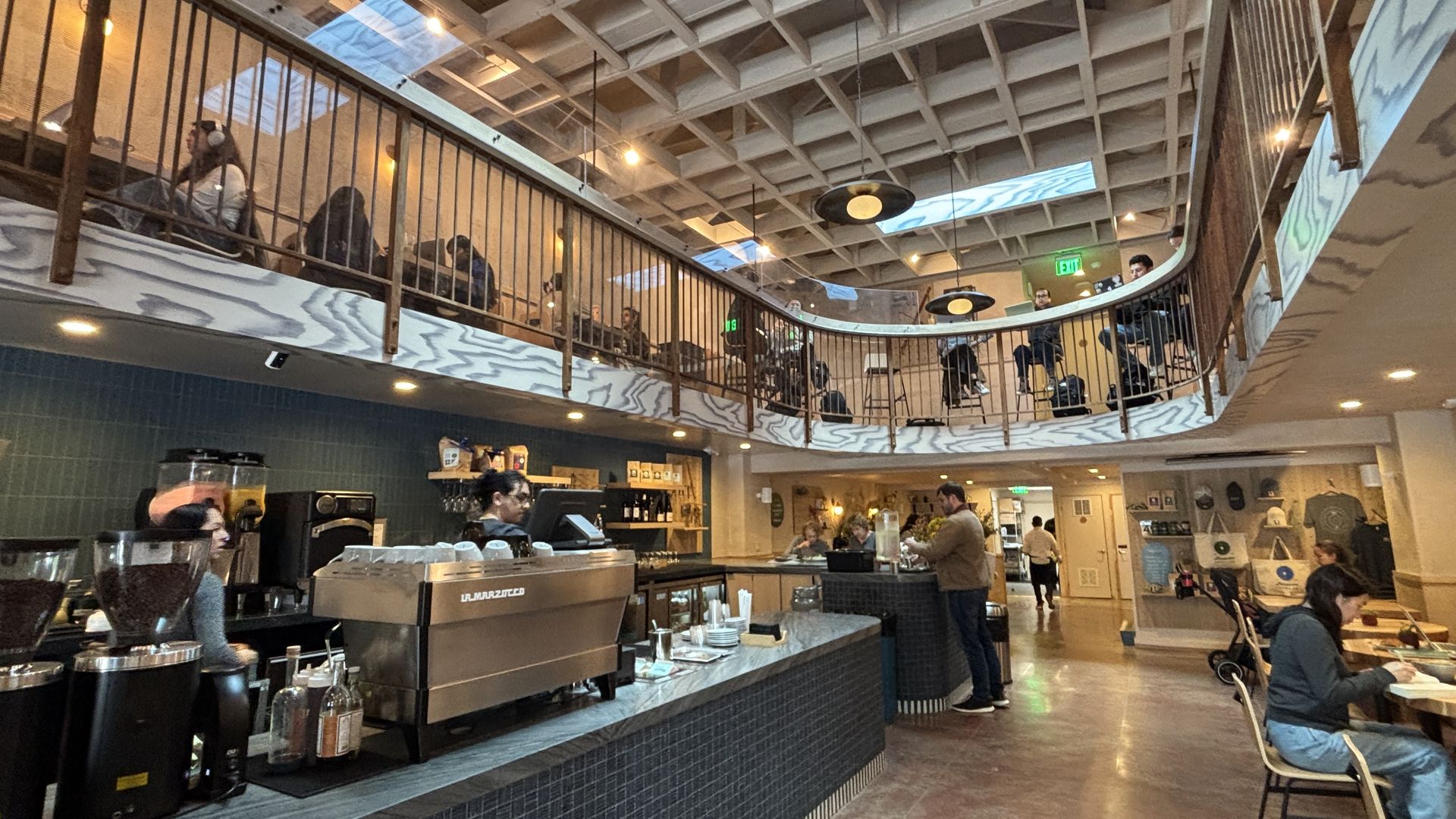 Cafe interior with a curved two-level balcony and wooden railing. Exposed beams, teal wall tiles, and warm lighting. People sit on both levels as a barista operates a large espresso machine at the counter.