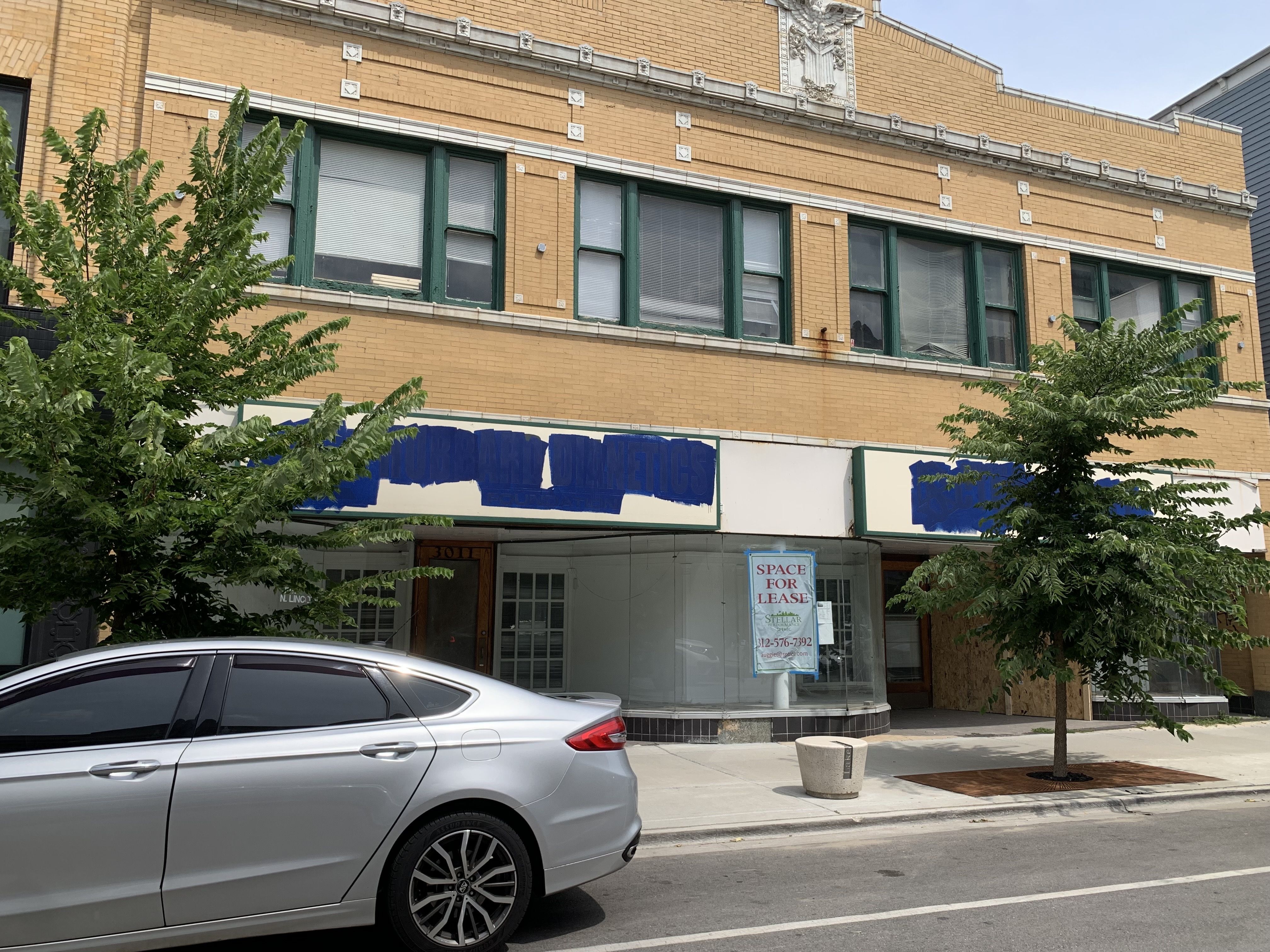 Yellow brick commercial building with green-framed windows, two small trees on the sidewalk, a silver car parked on the street, and empty storefronts with a "Space for Lease" sign.