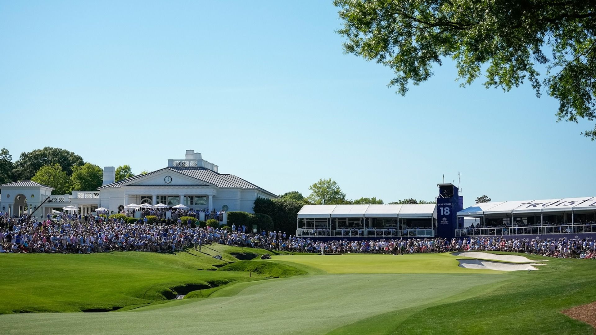 Sunny golf tournament scene: a large crowd along the manicured green, white clubhouse with a clock, white event tents, and a blue sky. Tree branches frame the top right.