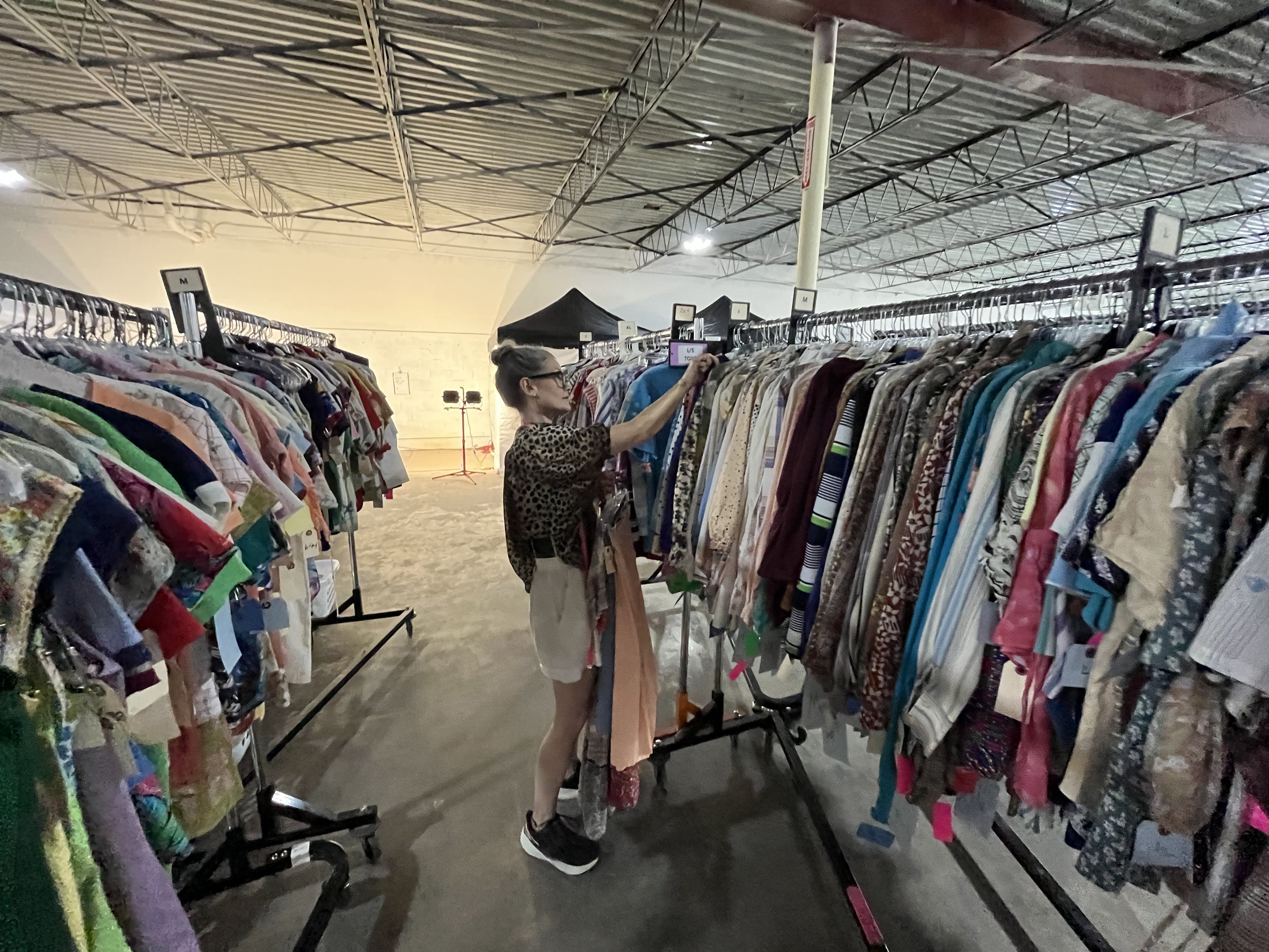 A woman in a leopard-print top and light shorts browses garments on long racks in a warehouse thrift store. Industrial ceiling, bright lights, and a small black canopy in the background.