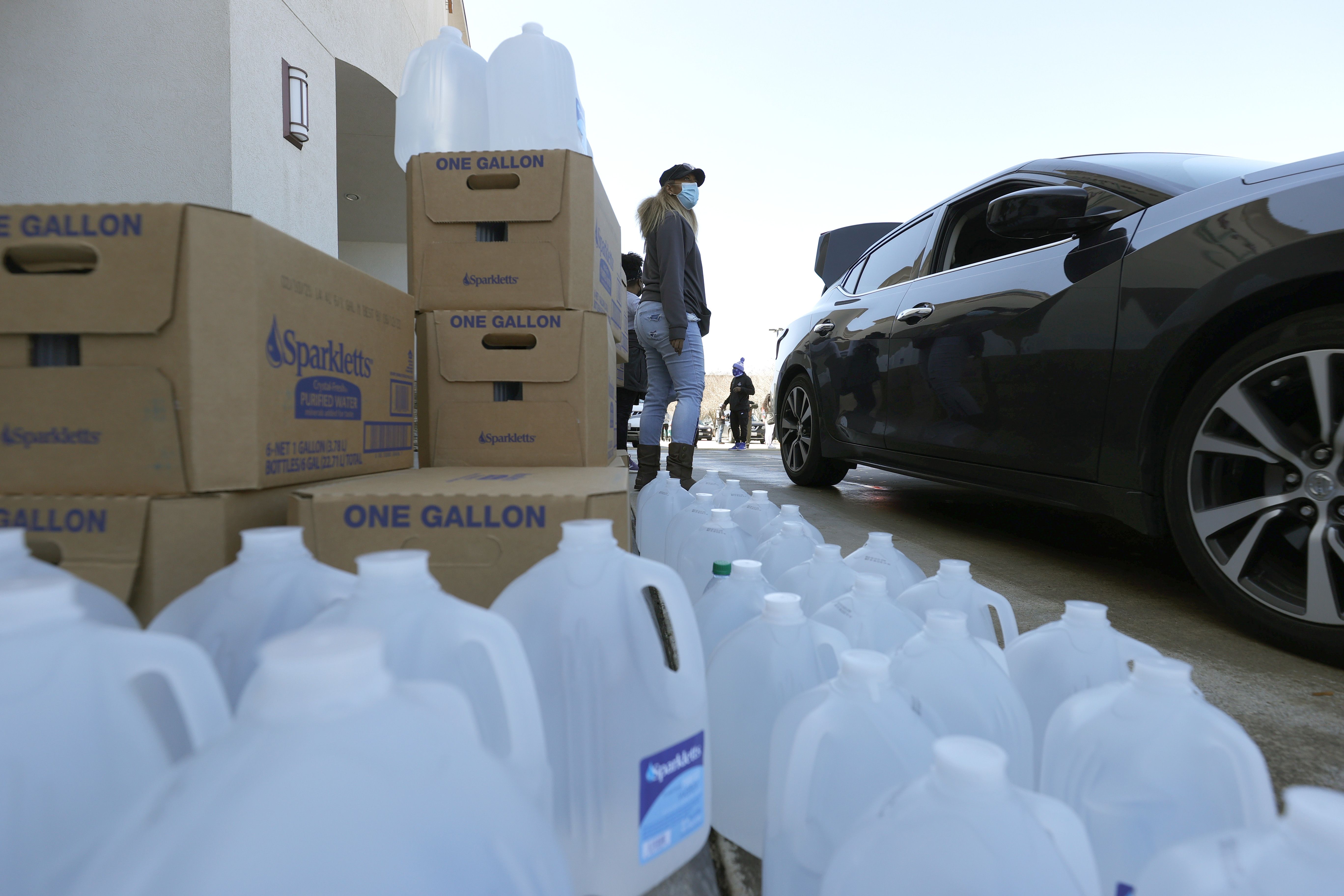 Dozens of gallons of water sit on the ground next to a car and cardboard boxes