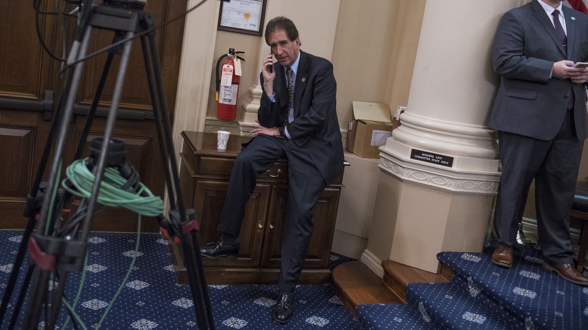 Two men in a government office: one in a dark suit sits and talks on a phone; another in gray stands by a column with the American flag behind. Foreground shows a camera on a tripod and a fire extinguisher.