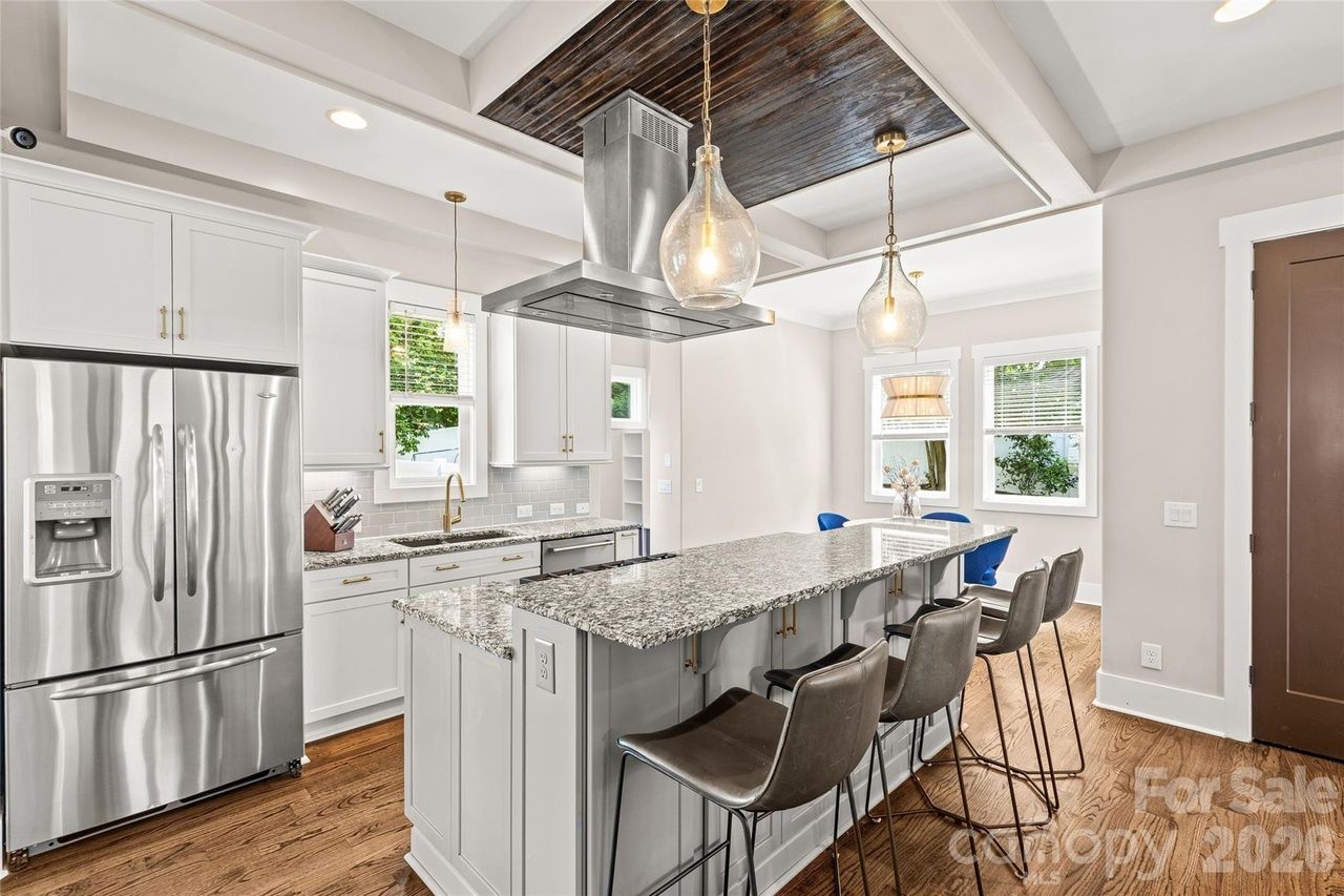 Bright kitchen with white cabinets, granite island, gray stools, stainless fridge. Brass hardware, pendant lights, dark wood ceiling inset, hardwood floors; dining area with blue chairs by windows.