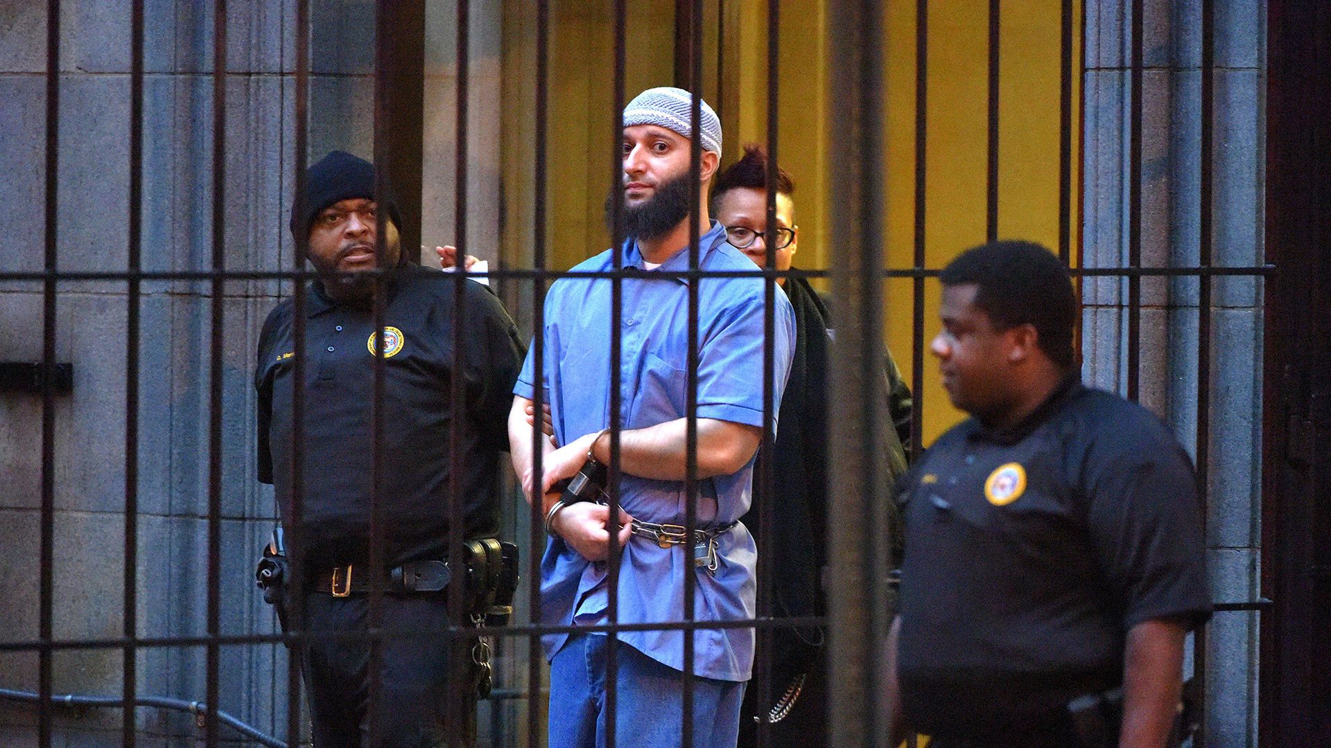Photo of Adnan Syed in a blue prison jumpsuit being escorted behind bars by guards