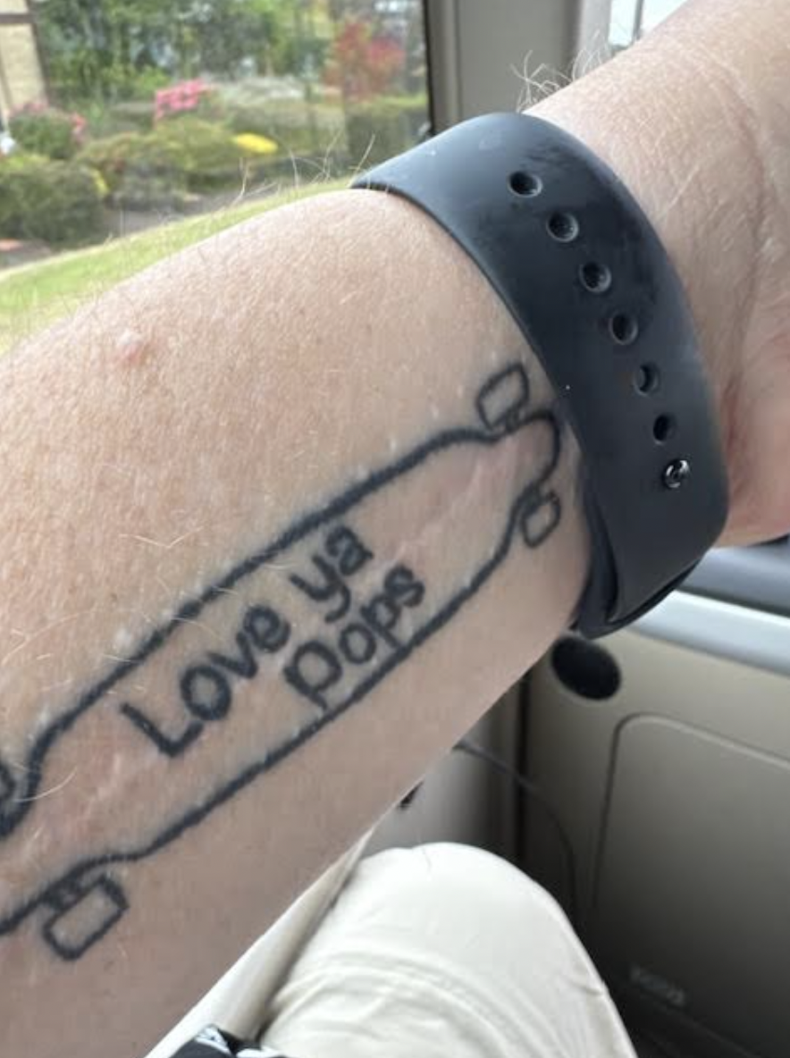 A tattoo on a man's forearm that says "Love ya, pops" on a longboard