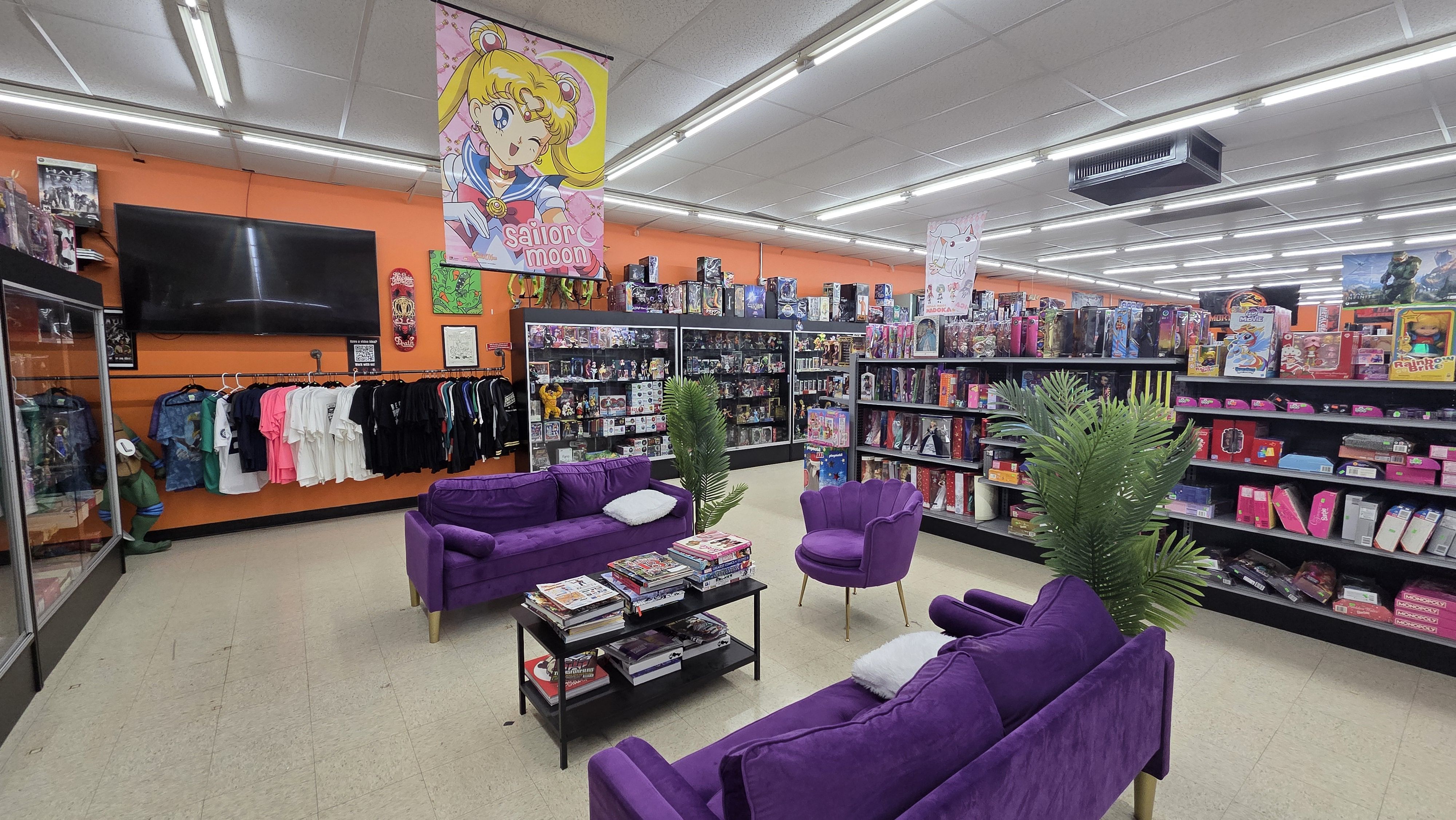 Bright store interior with orange walls, purple sofas and chair, shelves filled with toys and collectibles, a hanging Sailor Moon banner, and a TV mounted on the wall.
