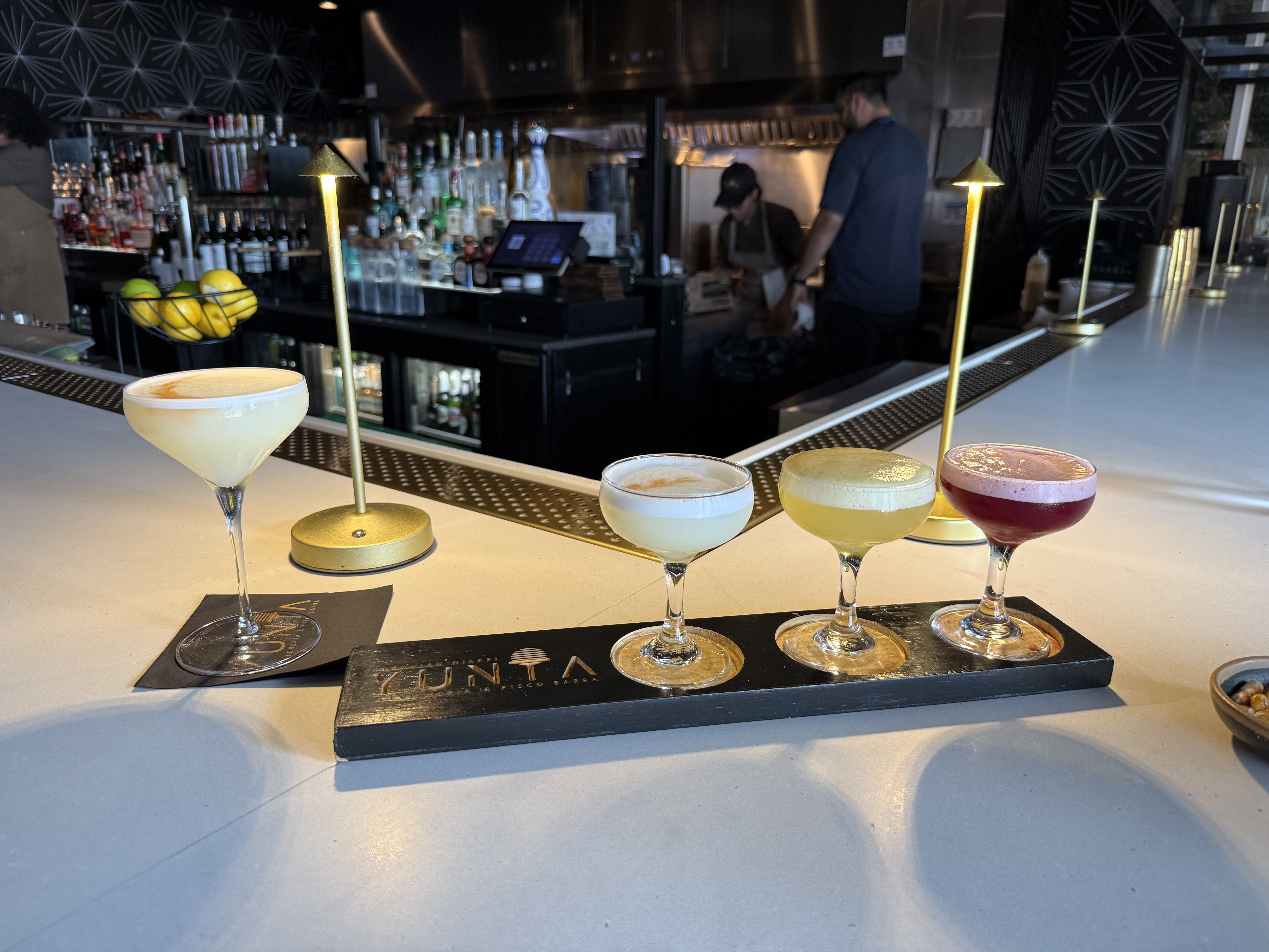 Five colorful cocktails line a marble bar in the foreground, with gold straws and a lemon basket nearby. In the background, a busy bar with bottles and two staff against a geometric patterned wall.
