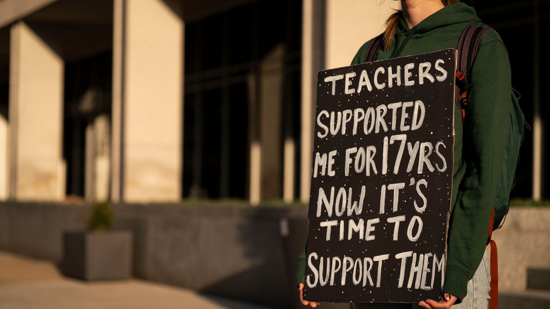 A person wearing a green hoodie stands holding a sign that reads "teachers supported me for 17 yrs now it's time to support them"