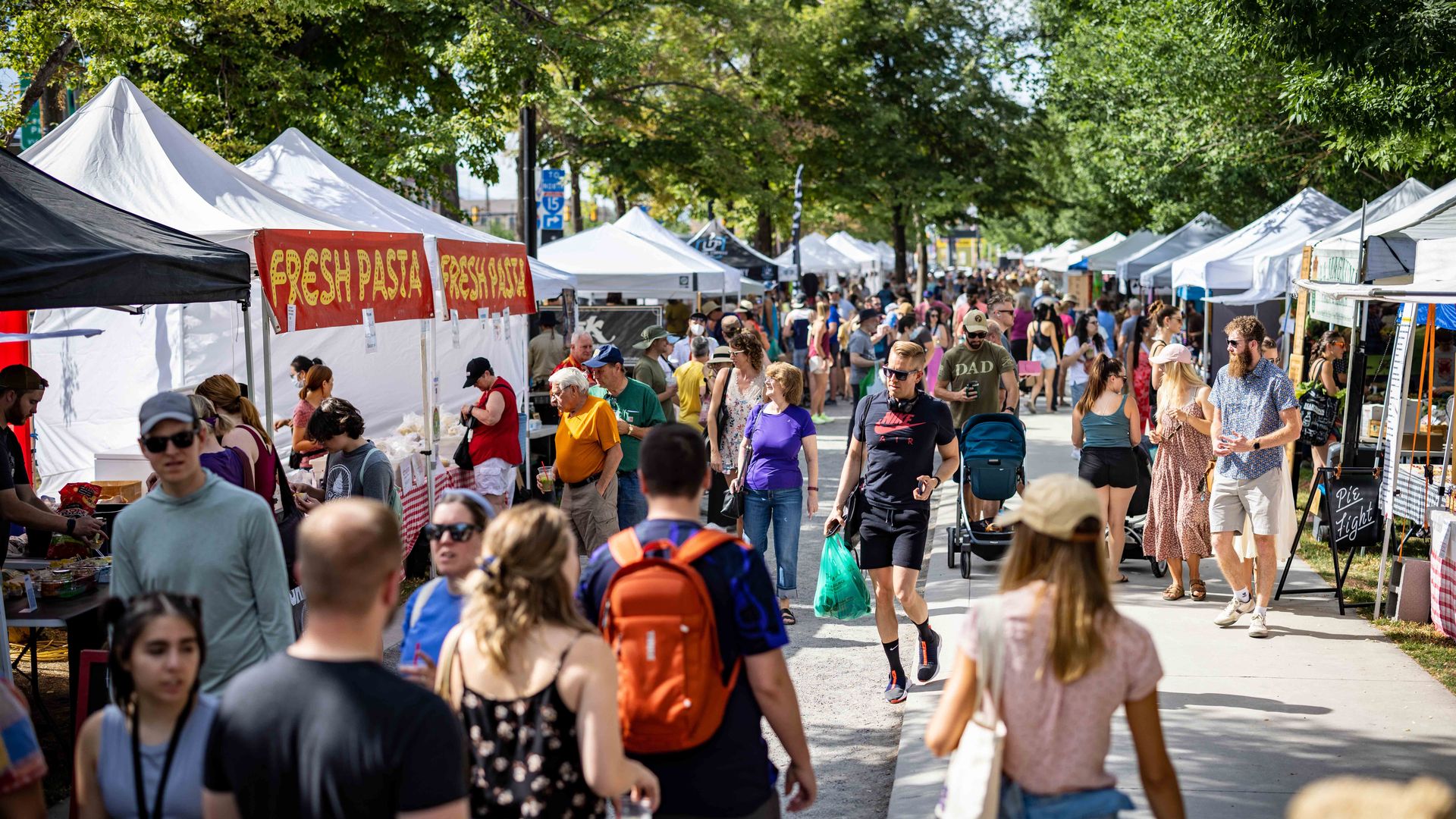 A busy street market with white vendor tents under leafy trees; crowds walk along the lane, many with drinks or strollers, while stalls serve food, including a red banner reading FRESH PASTA.