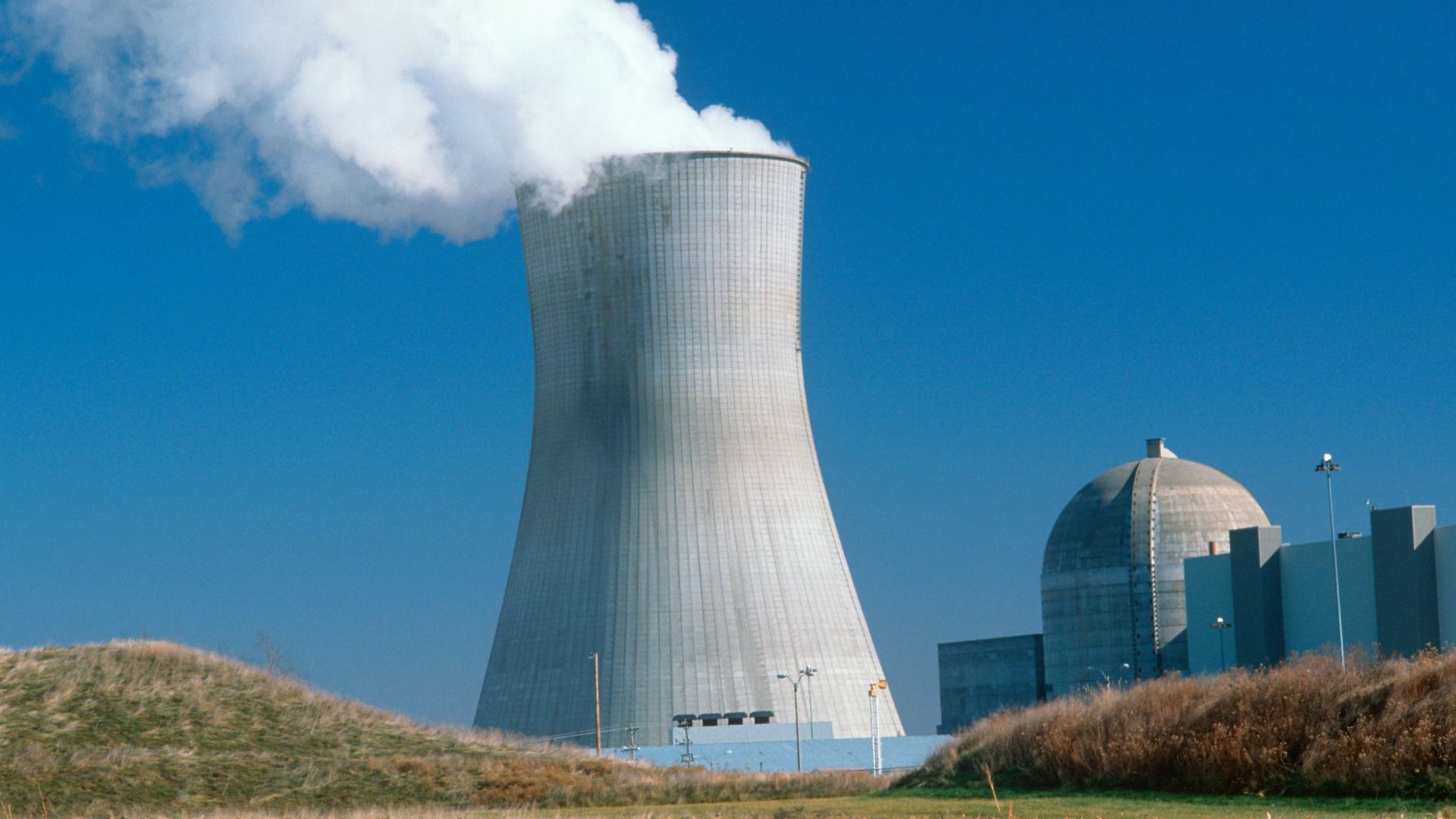 Photo shows a large cooling tower with steam coming out next to a facility with a dome, part of the Callaway Energy Center nuclear plant, with a blue sky and grassy hills in the foreground.