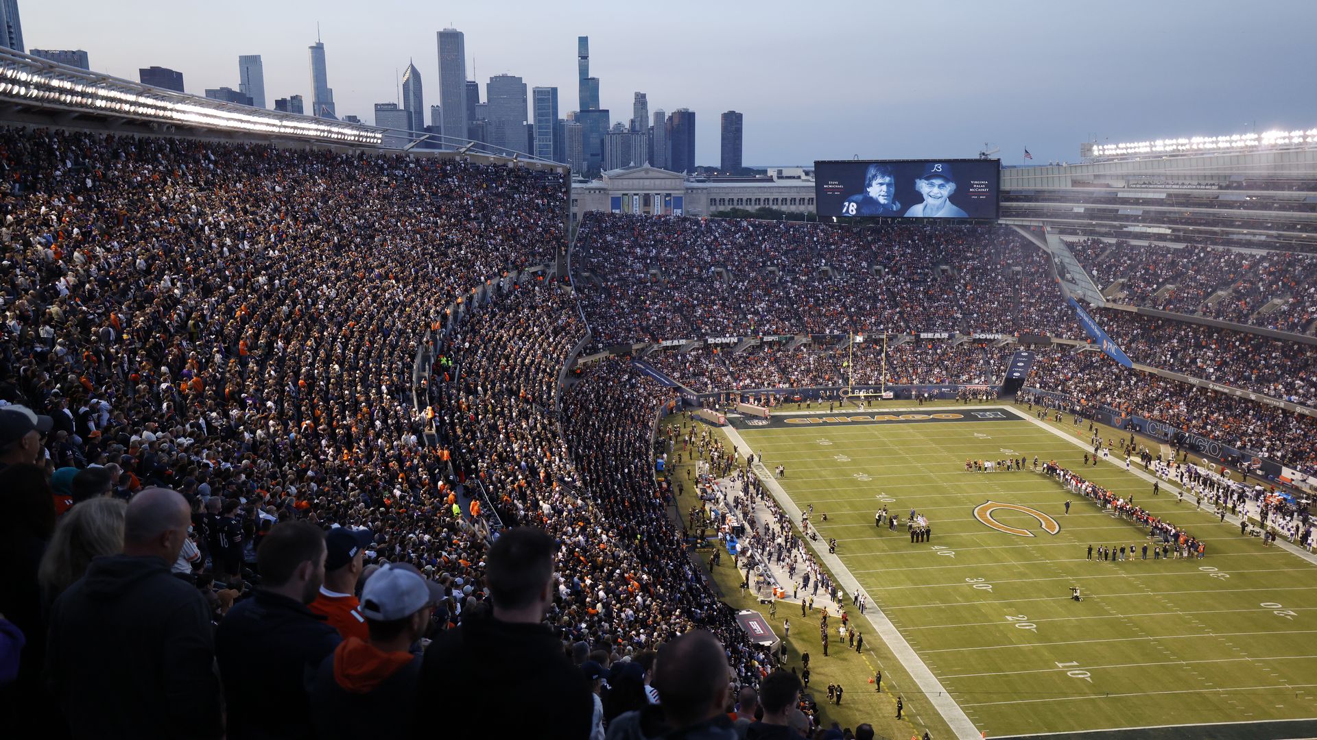 A packed Chicago Bears football stadium at dusk with fans wearing orange and navy. The field is lit and a large screen shows black-and-white portraits. Chicago skyline in background.