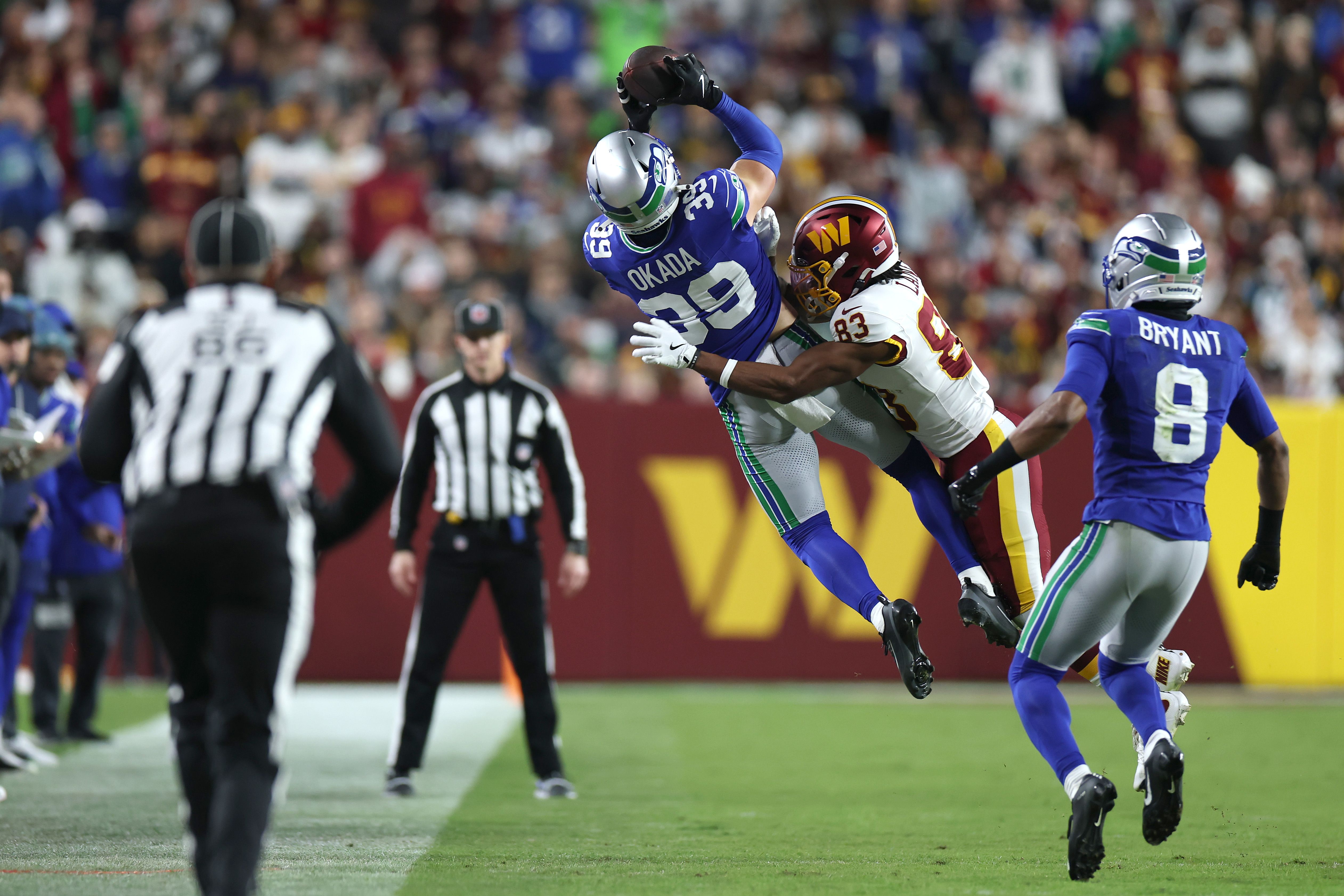 A player in a blue jersey with name Okada written at top jumps backward to catch a football as a player in a white jersey and maroon-brown helmet tries to stop him. Two referees and another blue-jerseyed player look on, with a blurry stadium crowd in the background.