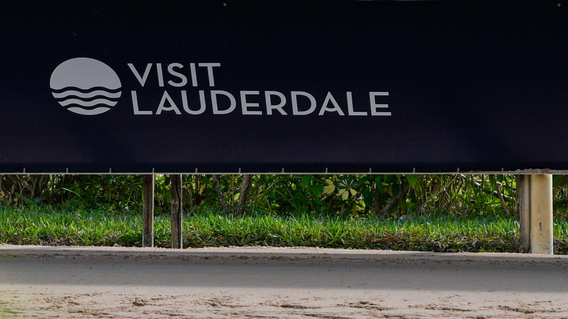 Black rectangular sign with Visit Lauderdale logo (in white writing) next to the side of the Gulfstream Park sidetrack. There is green shrubbery in the bottom foreground of the photo.