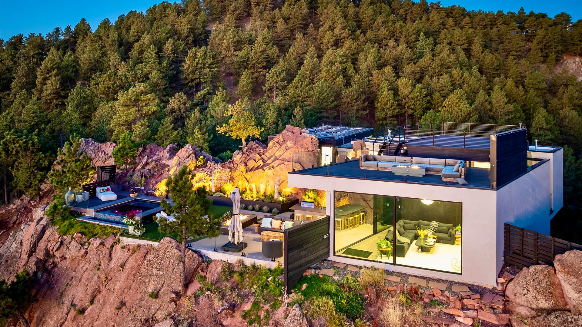 Modern house perched on rocky cliff surrounded by pine trees at dusk, featuring a rooftop lounge, glass walls, outdoor seating, hot tub, and warm ambient lighting.