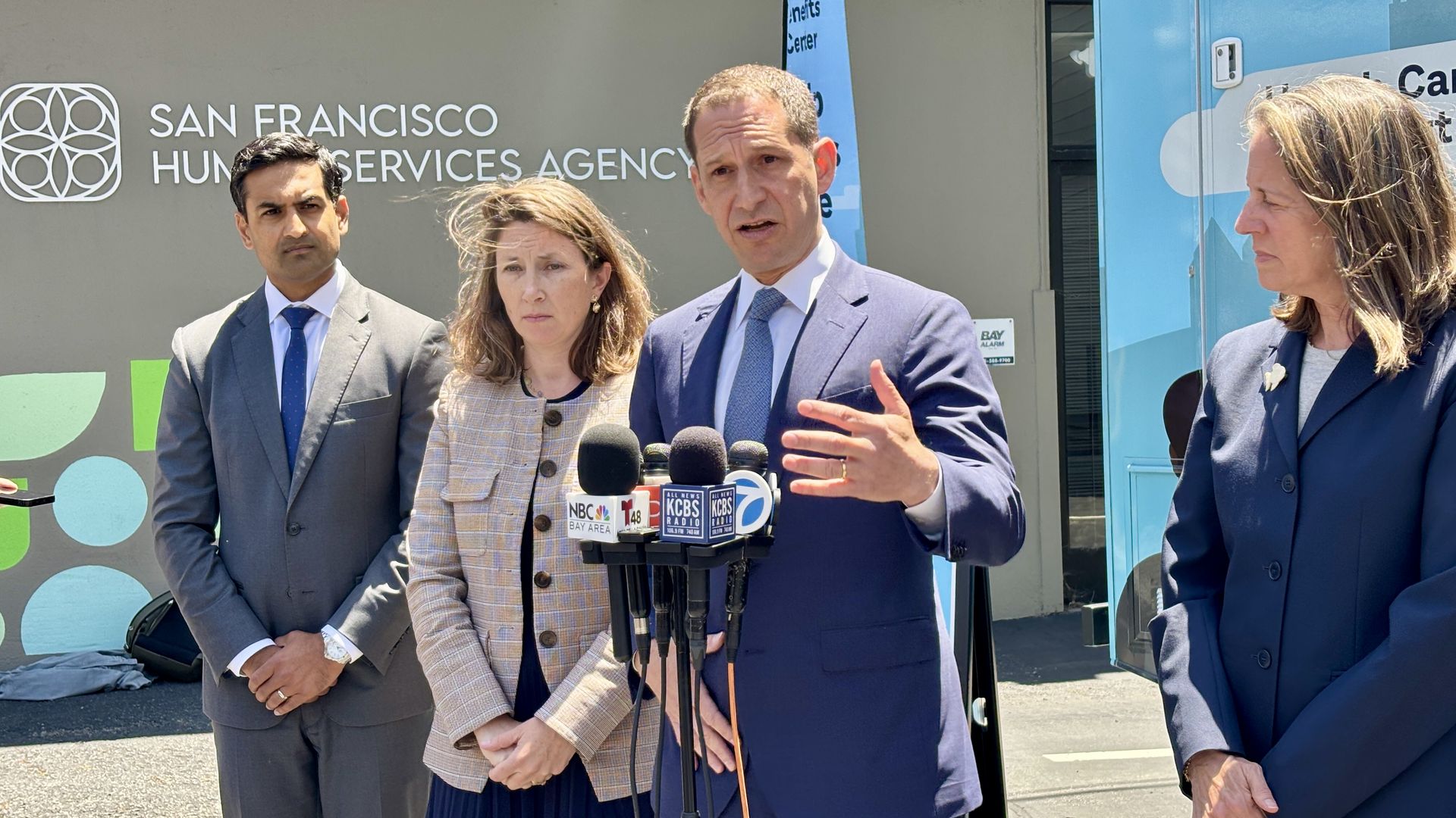Group of professionals at a press conference outside the San Francisco Human Services Agency. A man in a blue suit speaks into microphones, flanked by three officials, with a blue van behind.