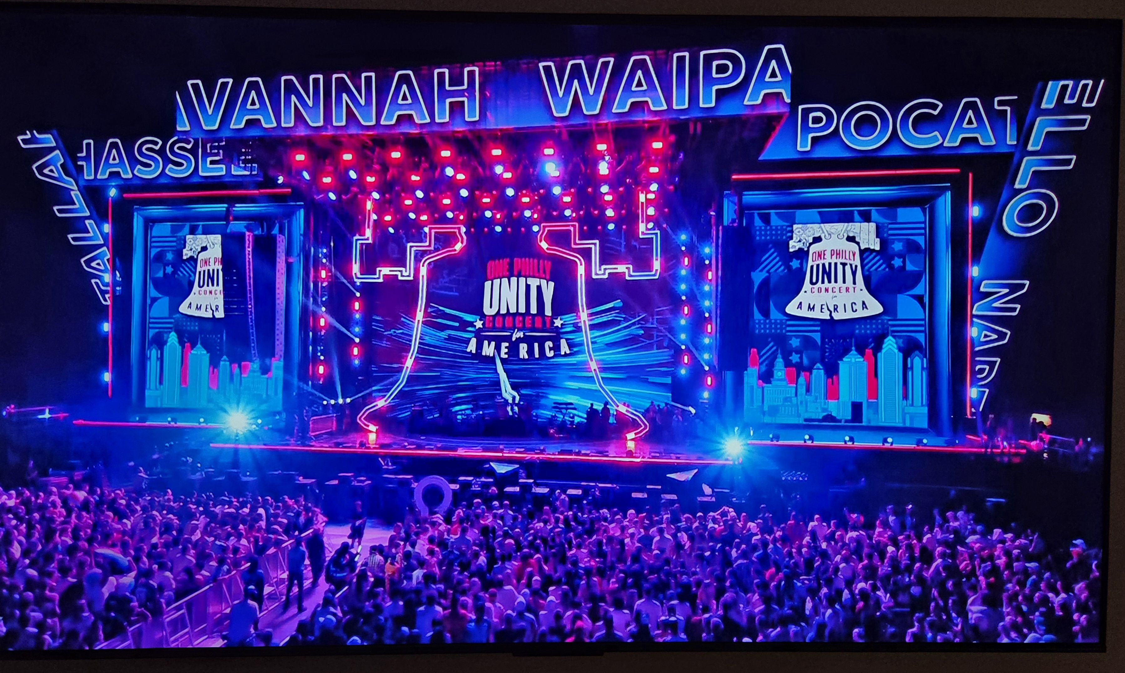 Neon-lit concert stage with blue and pink lights and a packed audience. Center screen reads "UNITY FOR AMERICA" with the "One Philly" logos, flanked by city skyline visuals.