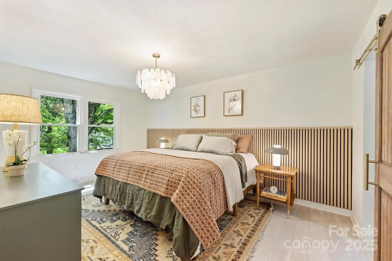Bright bedroom with large bed covered in patterned brown and beige bedding, two bedside tables with lamps, large window overlooking green trees, wooden slatted headboard, and chandelier.