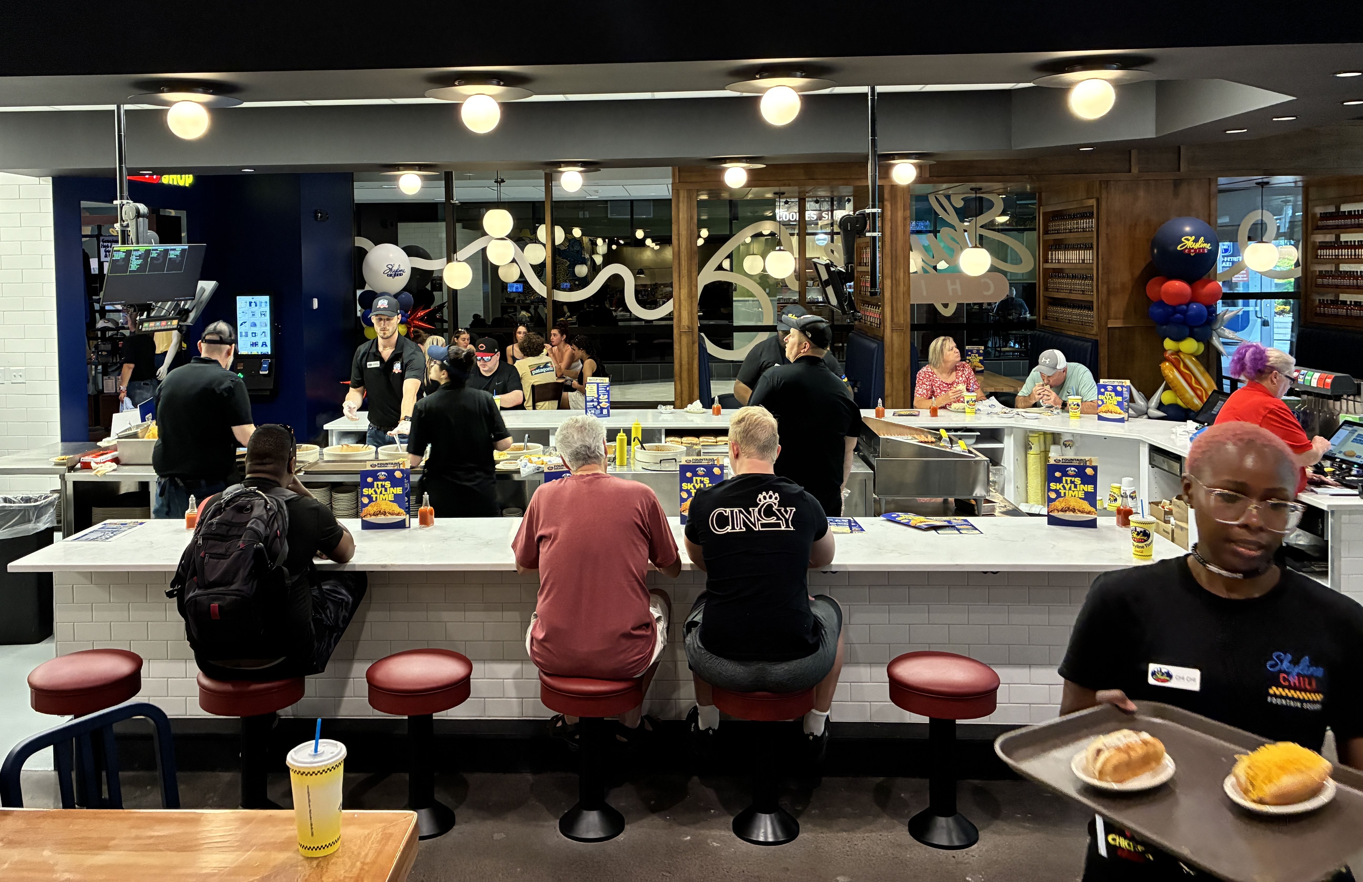 Busy Skyline Chili counter with diners on red stools at a white-tiled counter; staff in black shirts work under warm globe lights, with wood paneling, glass dividers, and balloons on the right.