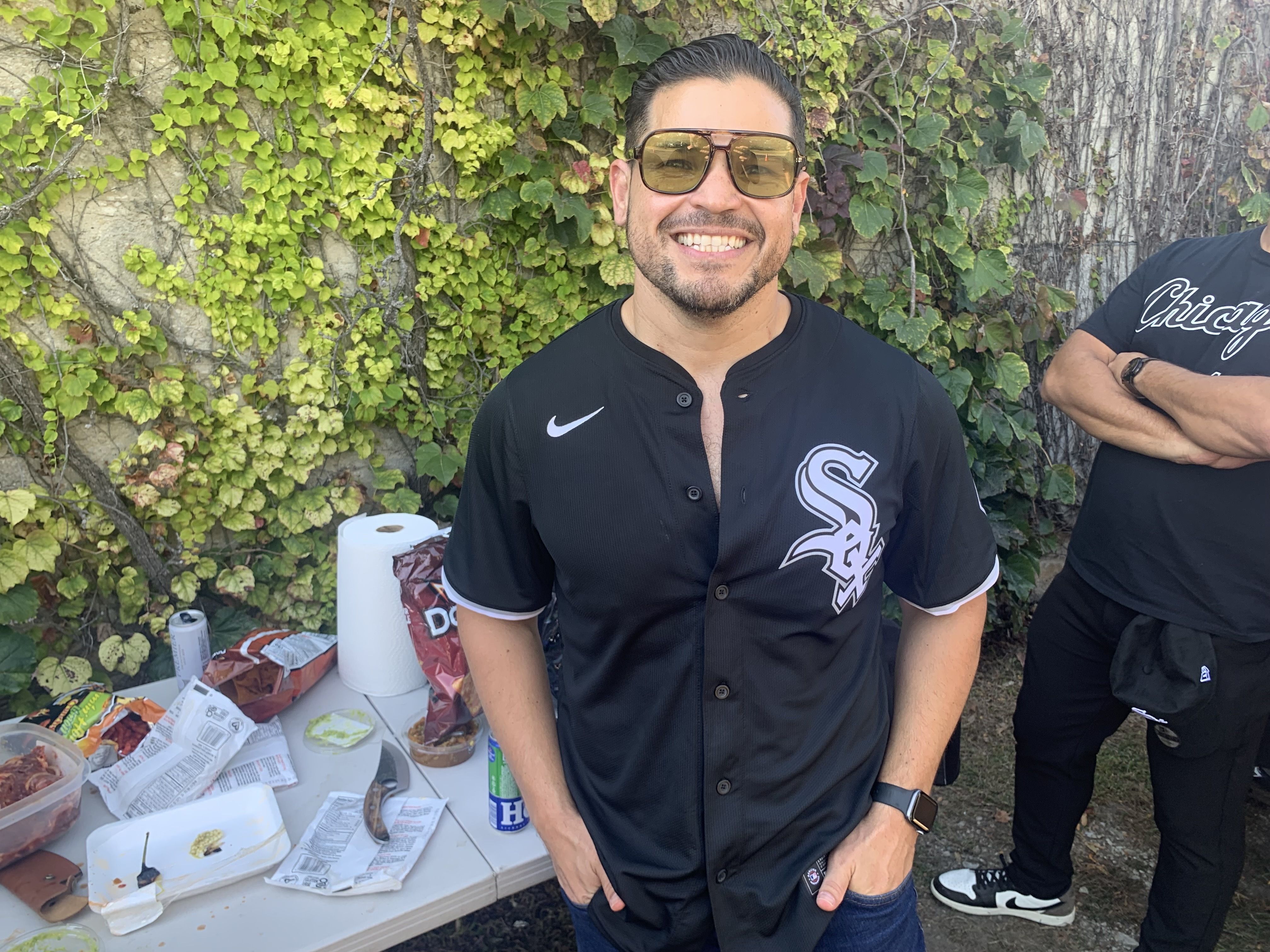 Smiling man in a black baseball-style shirt and brown sunglasses stands before an ivy-covered wall. A table with wrappers and drinks sits to his left, with a second person partly visible on the right.