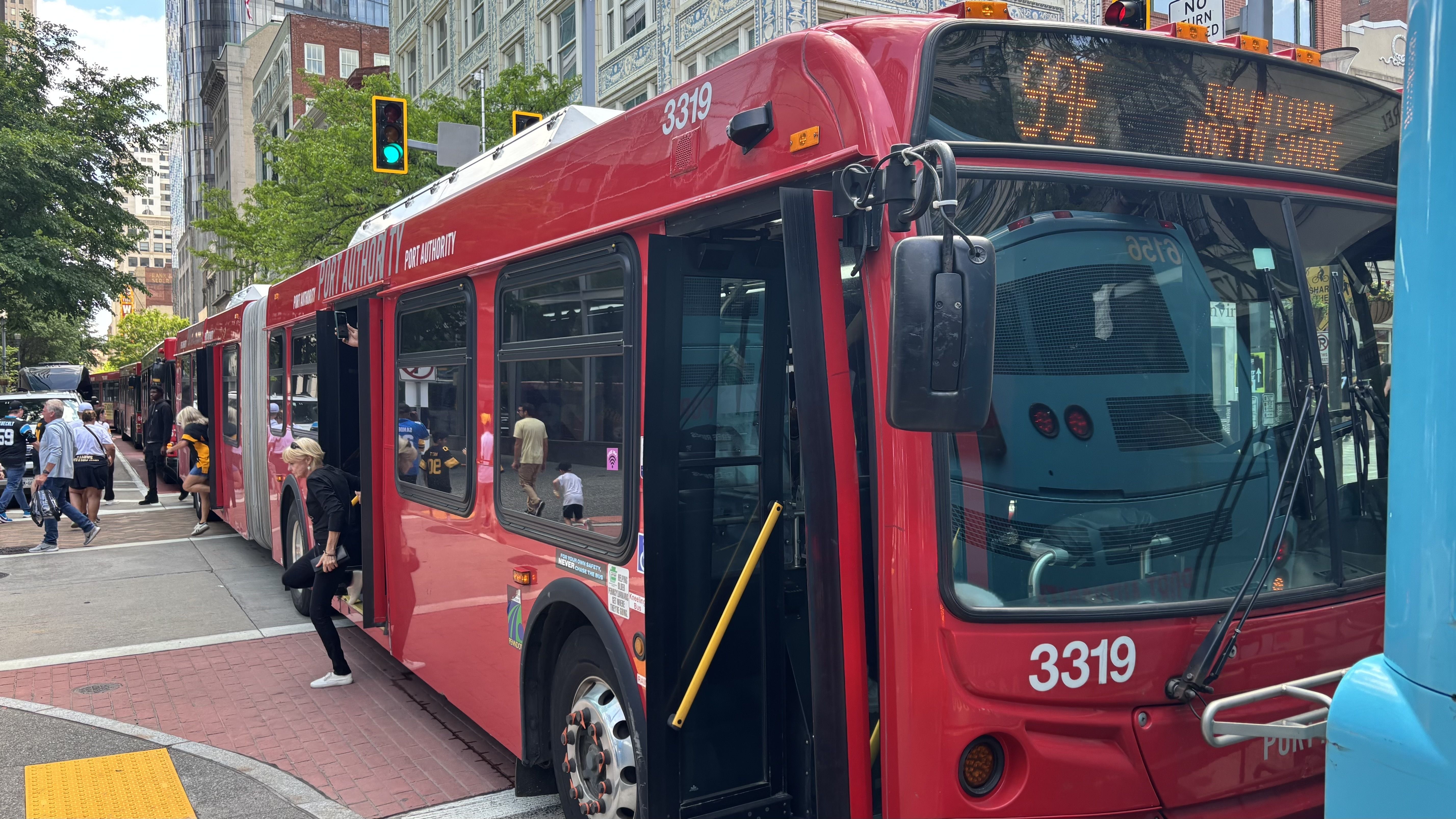 Red city bus with doors open, bus number 3319 and Port Authority on the side; people board on a busy urban street with tall buildings, green trees, and a green traffic signal.