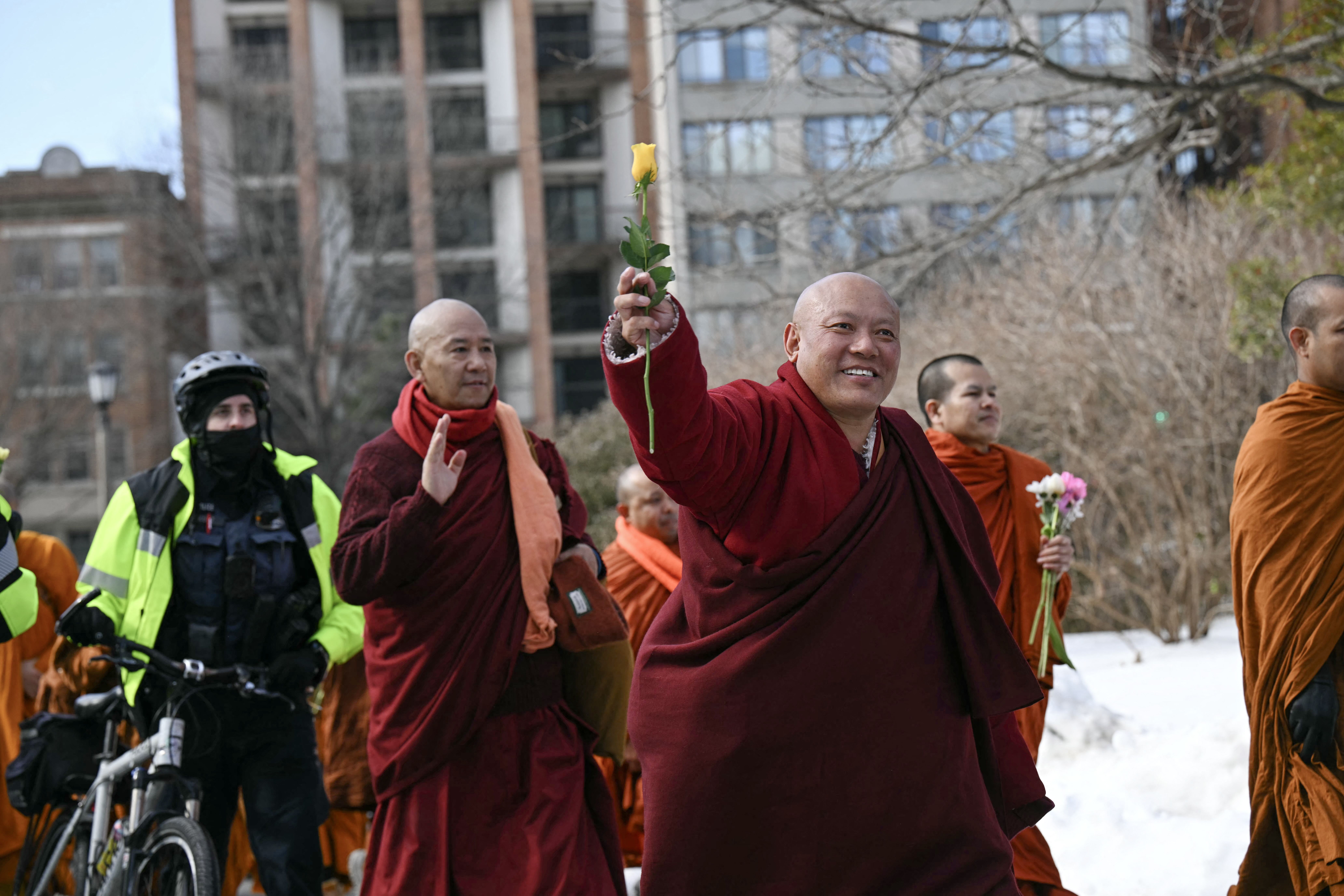 Group of Buddhist monks in red and orange robes walking outdoors in winter, one monk smiling and holding a yellow rose, accompanied by a police officer on a bicycle wearing bright yellow jacket.