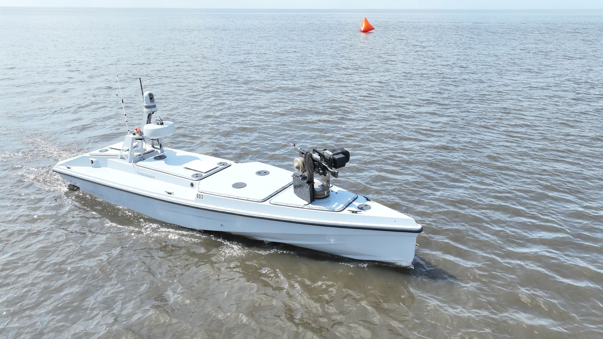 White unmanned naval boat with mounted machine gun and antennas cruising on calm water near an orange buoy.
