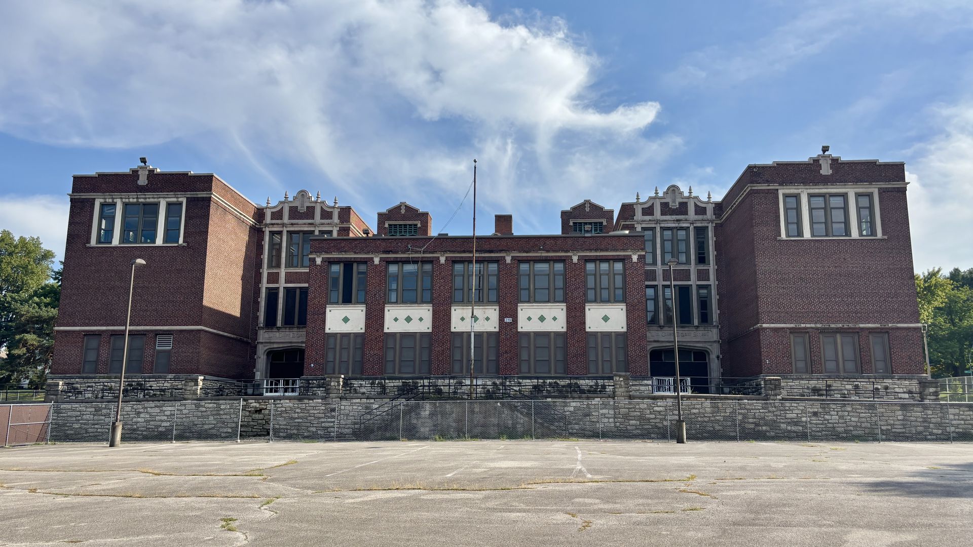 Wide view of a red brick, multi-wing building with white stone trim and large windows, set on a stone foundation behind a chain-link fence in an empty parking lot, under a blue sky with clouds.