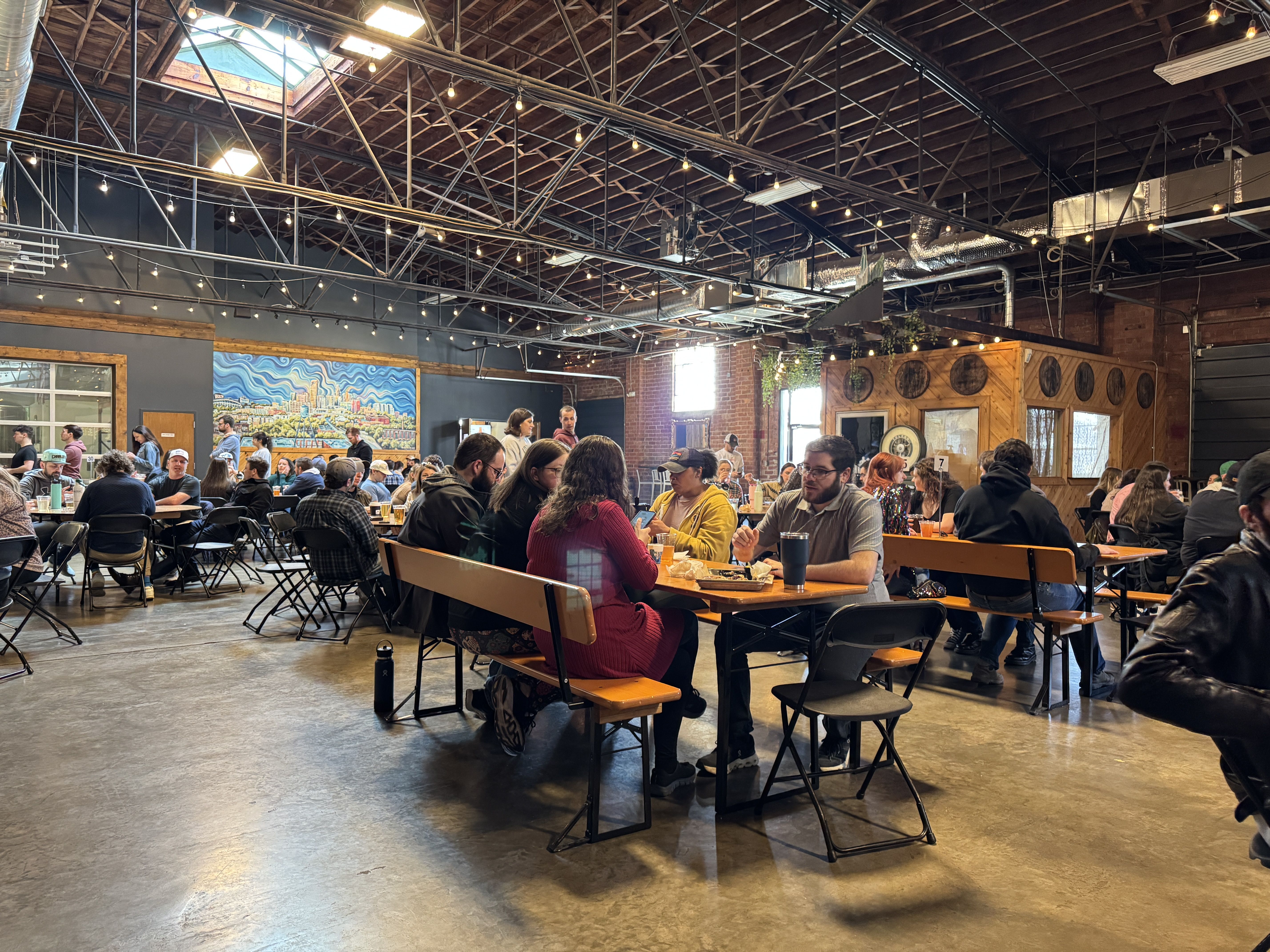People sitting and socializing at wooden tables with benches in a spacious indoor venue with high ceilings, string lights, a colorful city mural, and industrial decor.