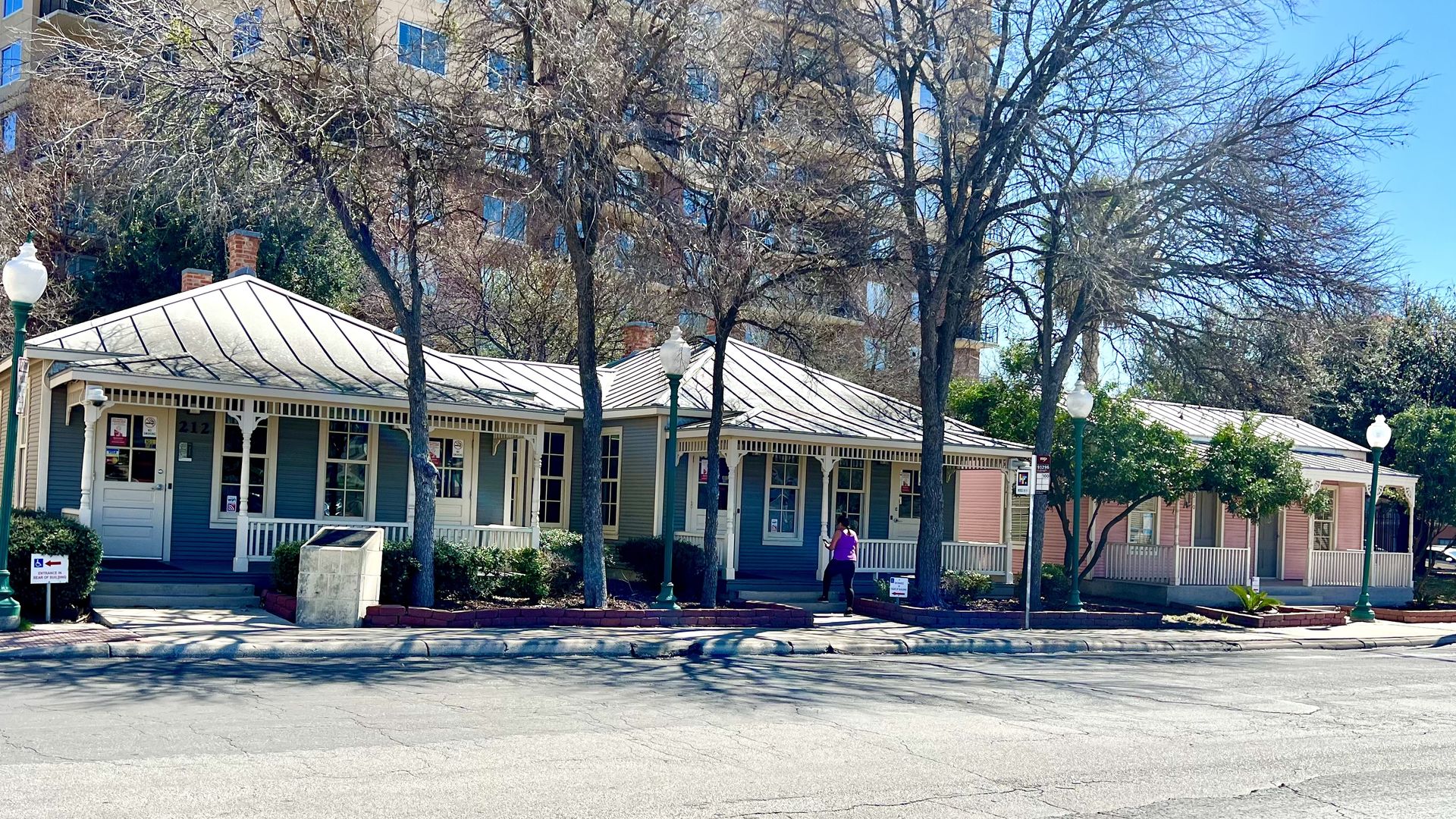 Row of blue and pink cottages with white porches and bare trees in front, set against a tall beige apartment building under a clear blue sky.