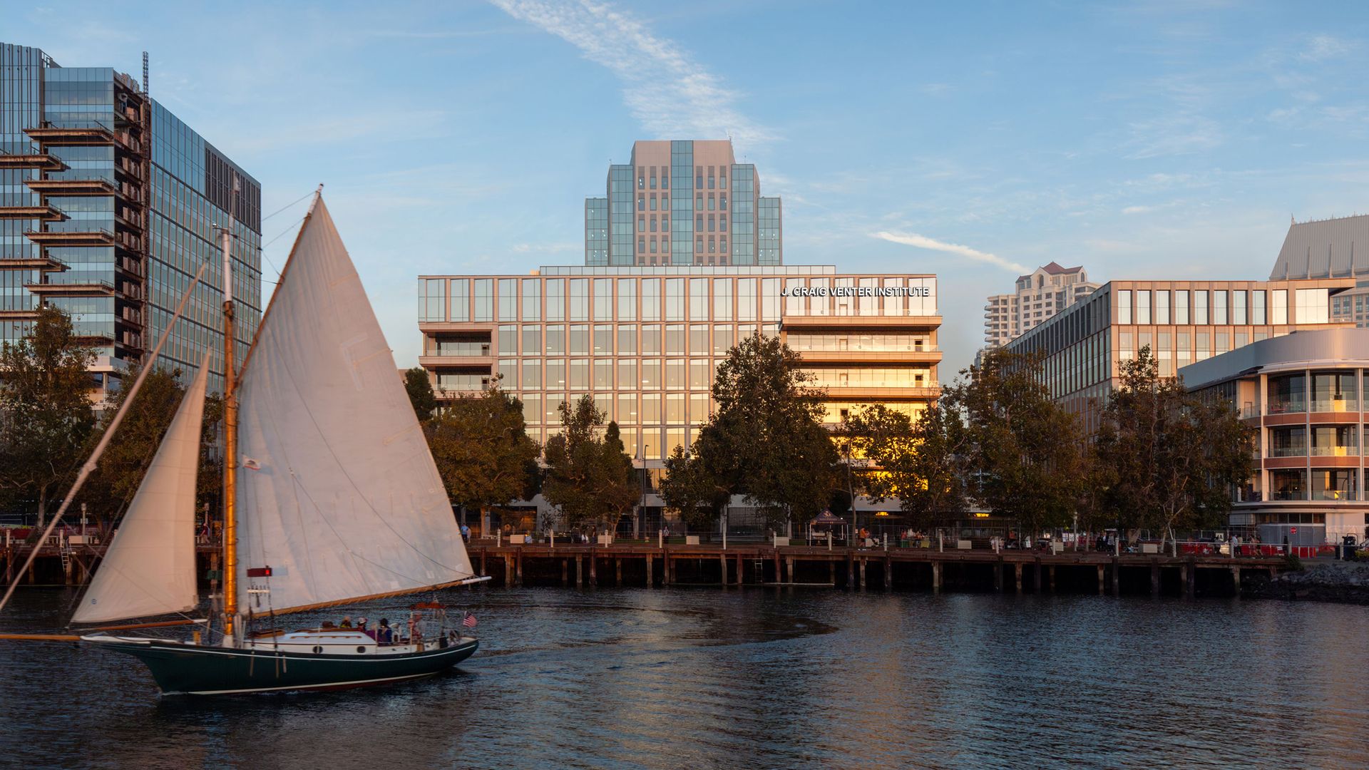 A rednering of an office building overlooking the downtown waterfront with J. Craig Venter on the top floor, with a sailboat in the water in the foreground and a dock along the shore 