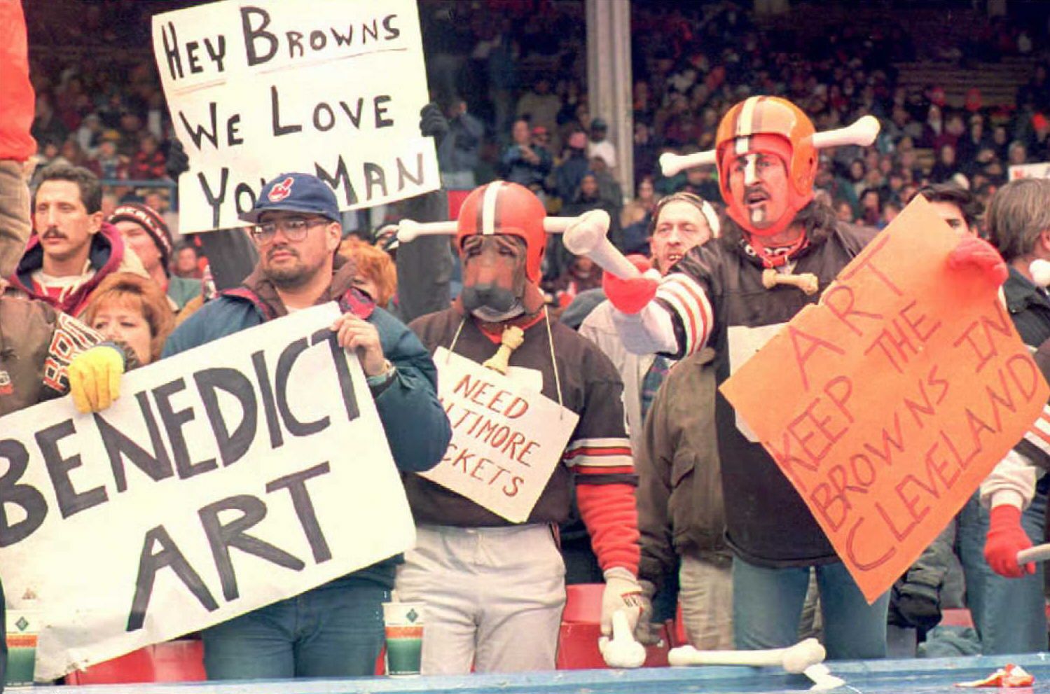Crowd of football fans at a stadium, many in costumes with oversized helmets, holding handmade signs like "Hey Browns We Love You Man" and "Benedict Art," with an orange sign visible on the right.