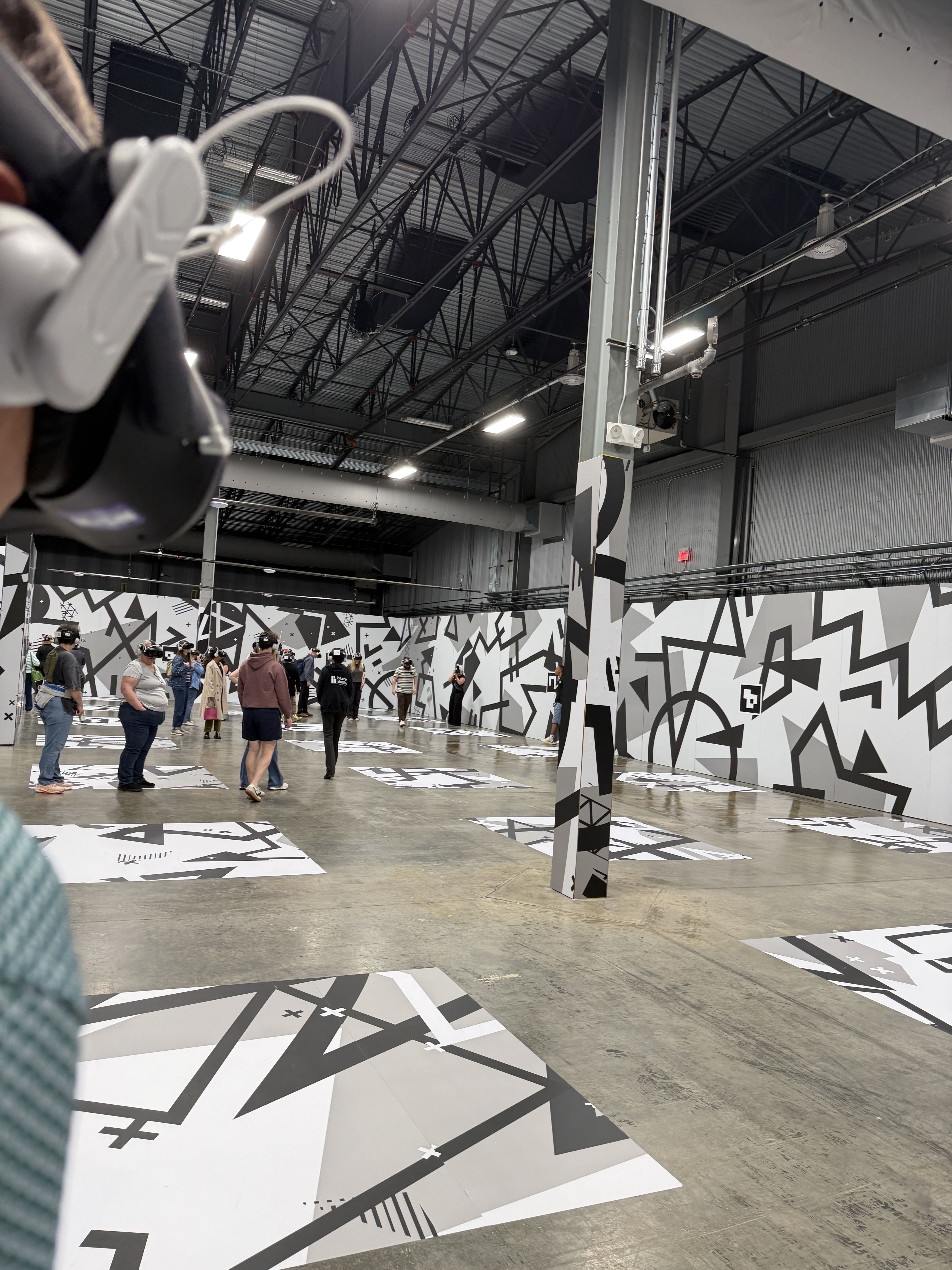 Industrial gallery with black-and-white geometric murals on walls and matching floor panels; a group of people observe the art. Exposed ceiling trusses and a tall striped pillar frame the space.