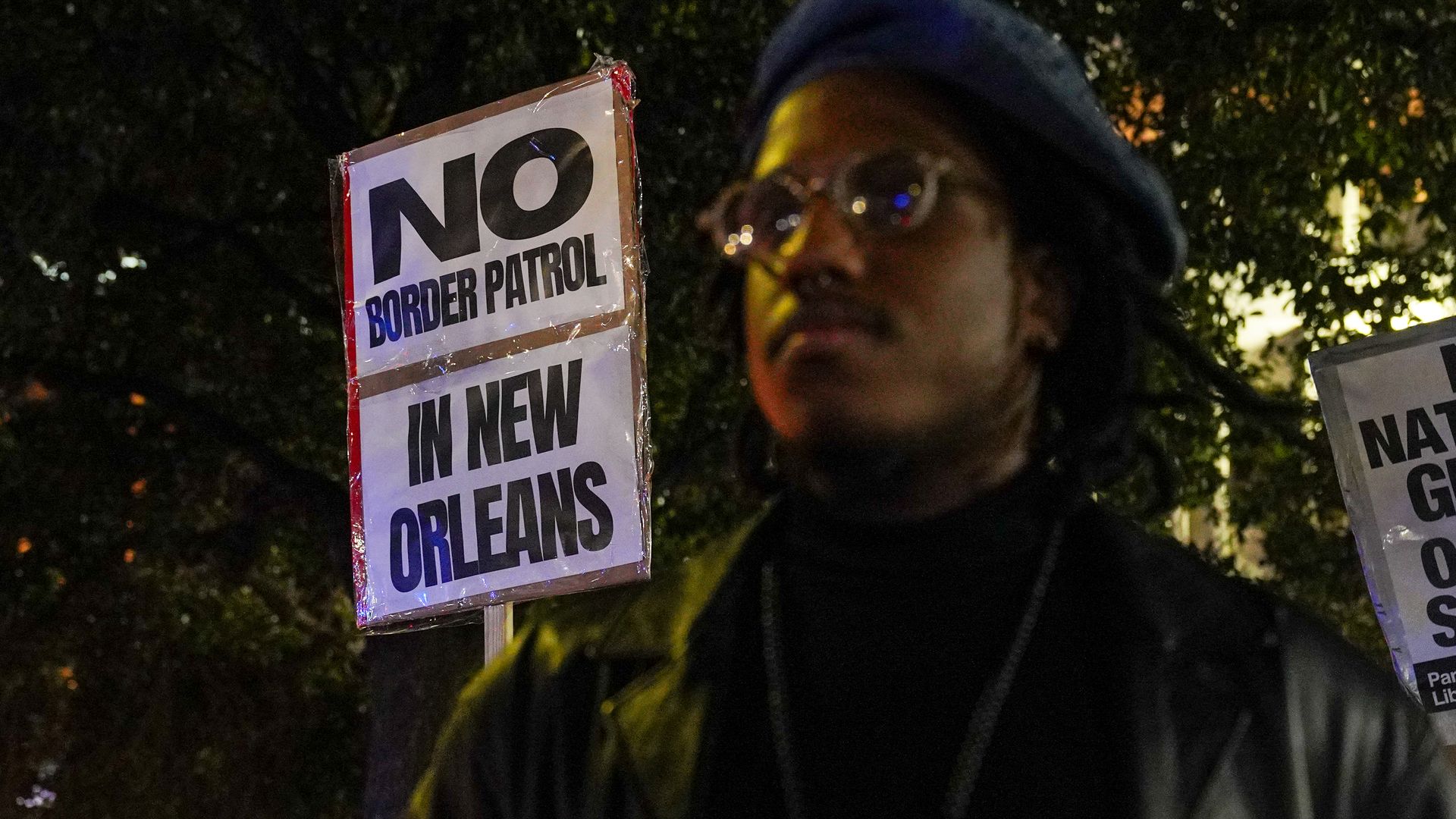 A person is photographed close-up wearing all black. Behind them, a protest sign reads "No Border Patrol in New Orleans."