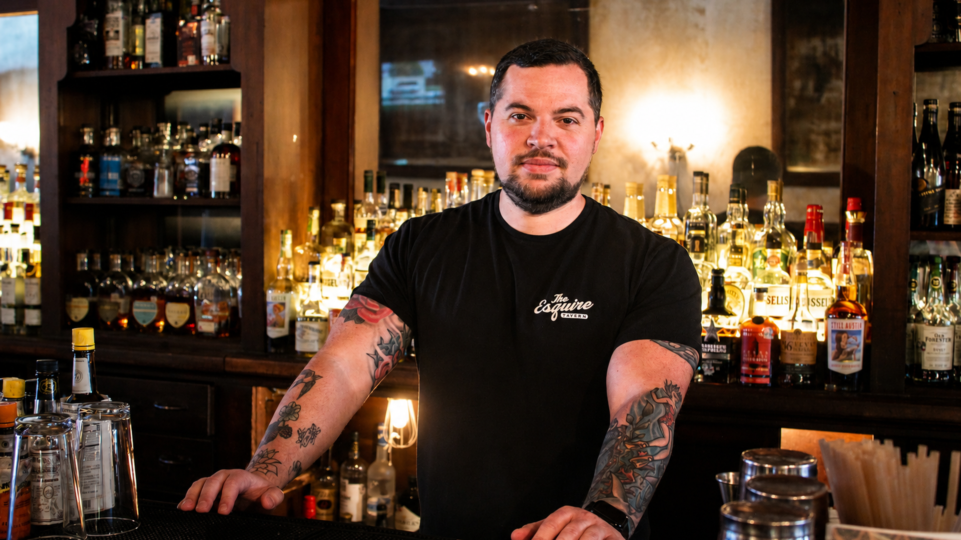 Tattooed bartender in a black T-shirt stands behind a well-lit bar, hands on the counter. Shelves of liquor bottles glow in the background, creating a warm, inviting tavern atmosphere.