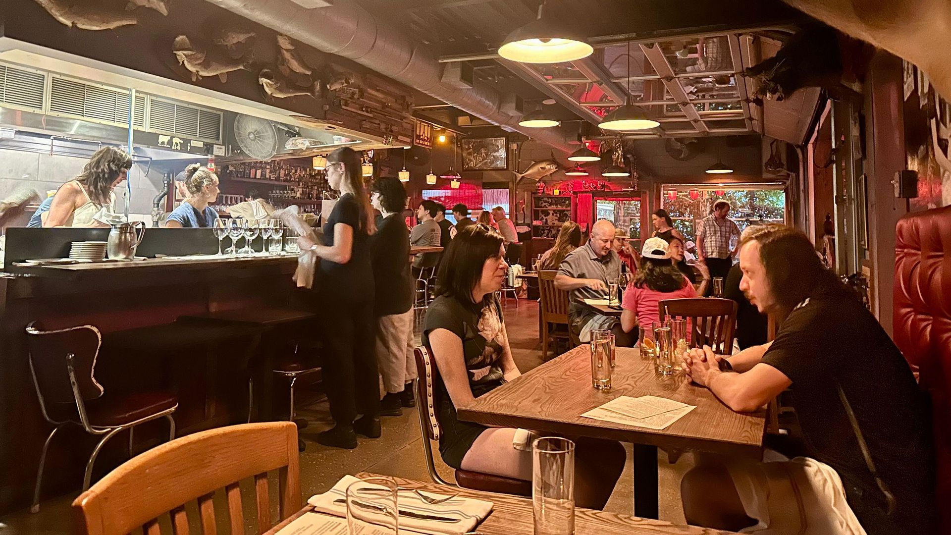 Busy restaurant interior with wooden tables, red cushioned booths, patrons dining and chatting, waitstaff busy near a bar with glassware, dim lighting, and fish mounted on walls and ceiling.