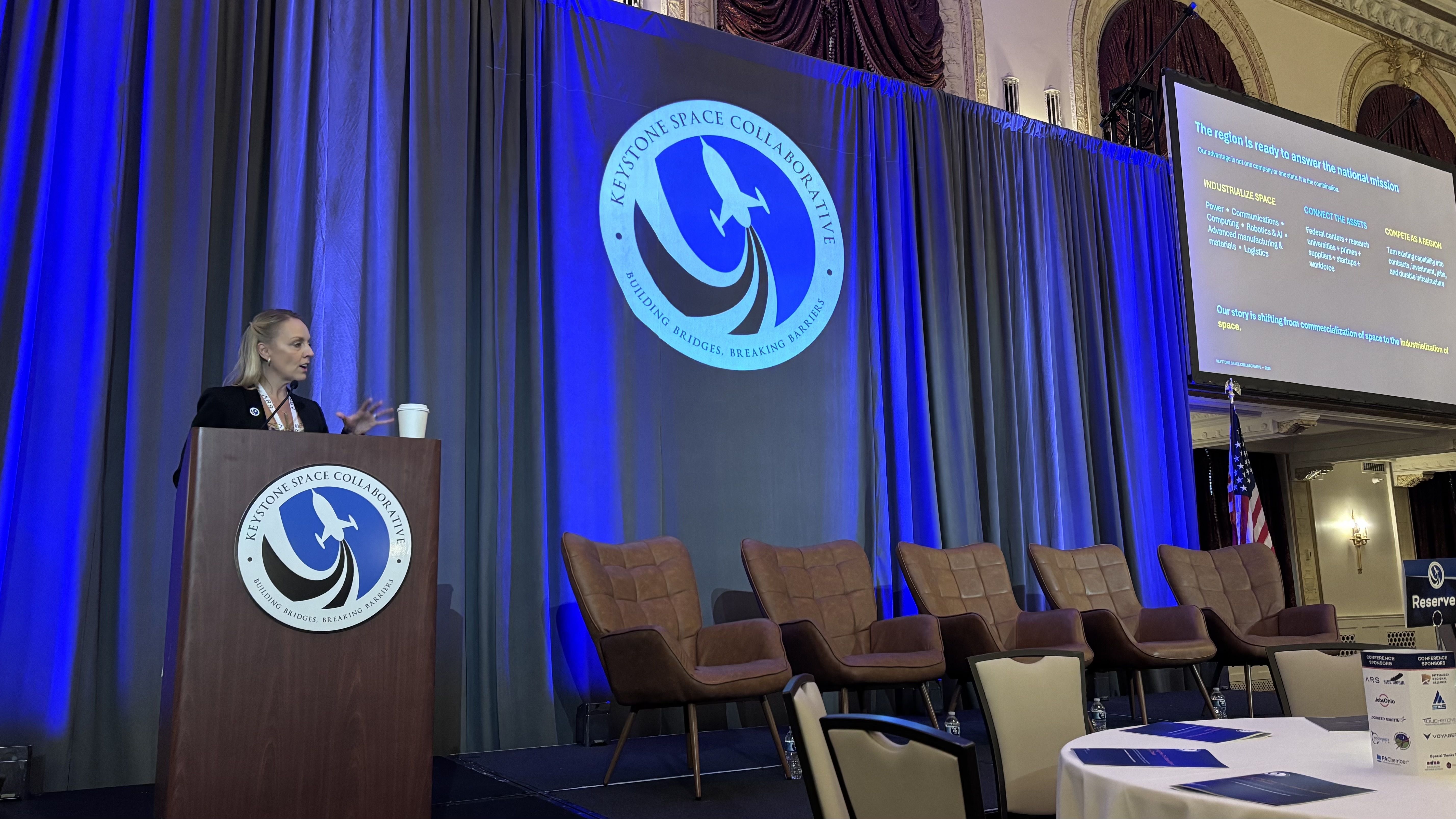 Speaker at a podium on a blue-lit conference stage, with a large circular Keystone Space Collaborative logo on the curtain and a projection screen to the right displaying slides; empty chairs in front.