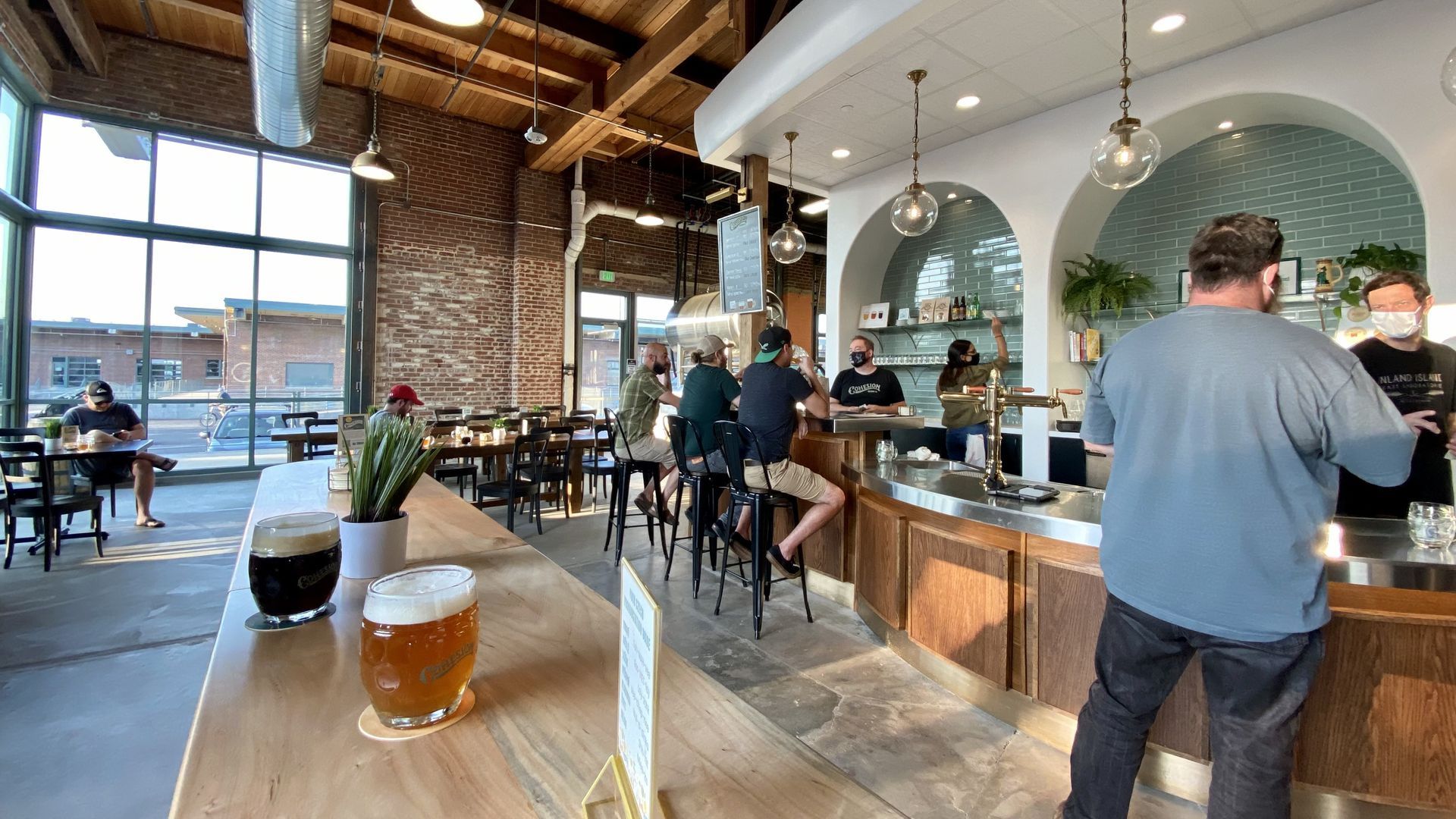 Two beers sit on a table at a Denver brewery with customers drinking in the background.