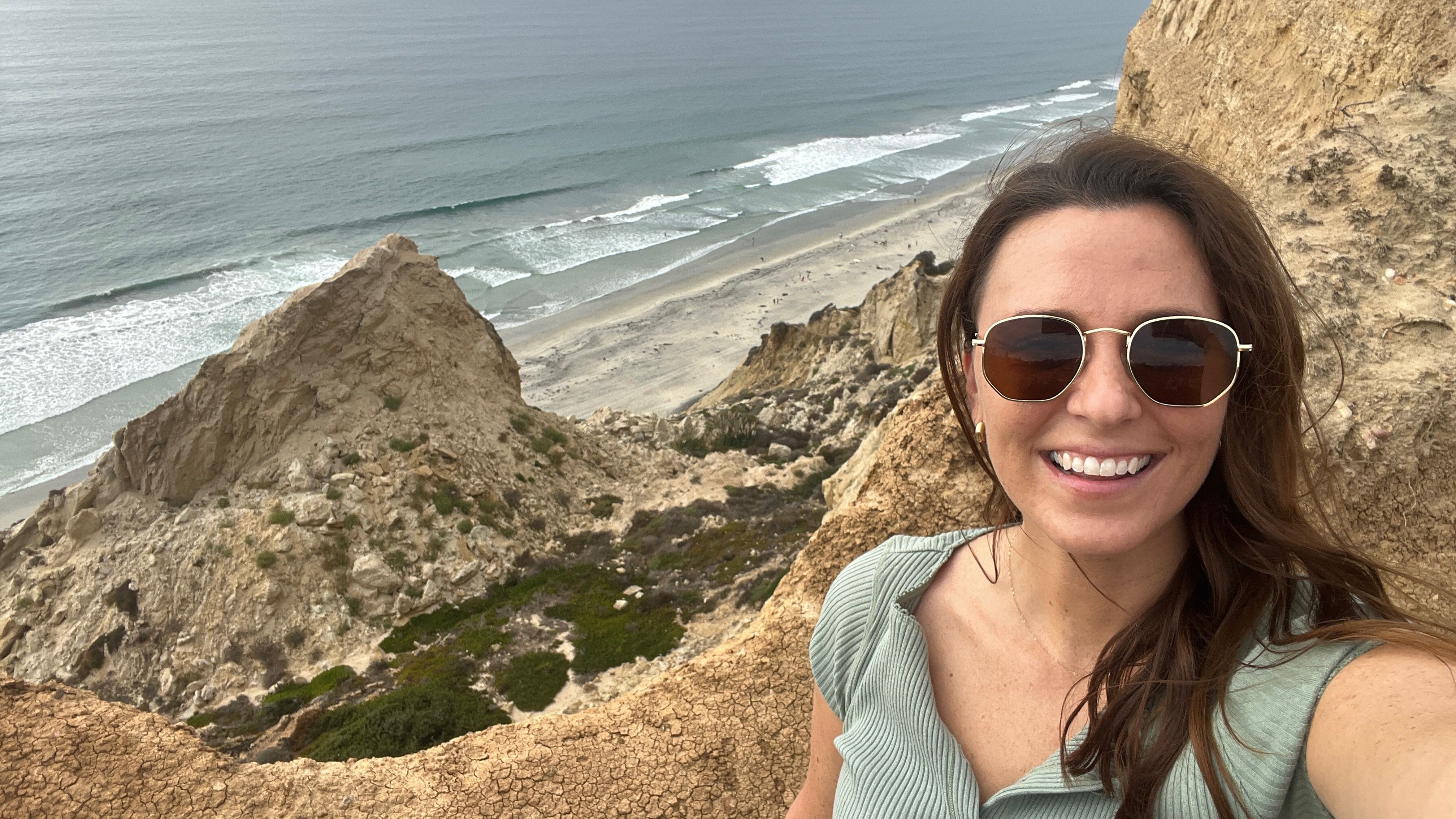 Woman wearing sunglasses and green top smiling, taking selfie on rocky cliff overlooking sandy beach and ocean waves on a cloudy day.