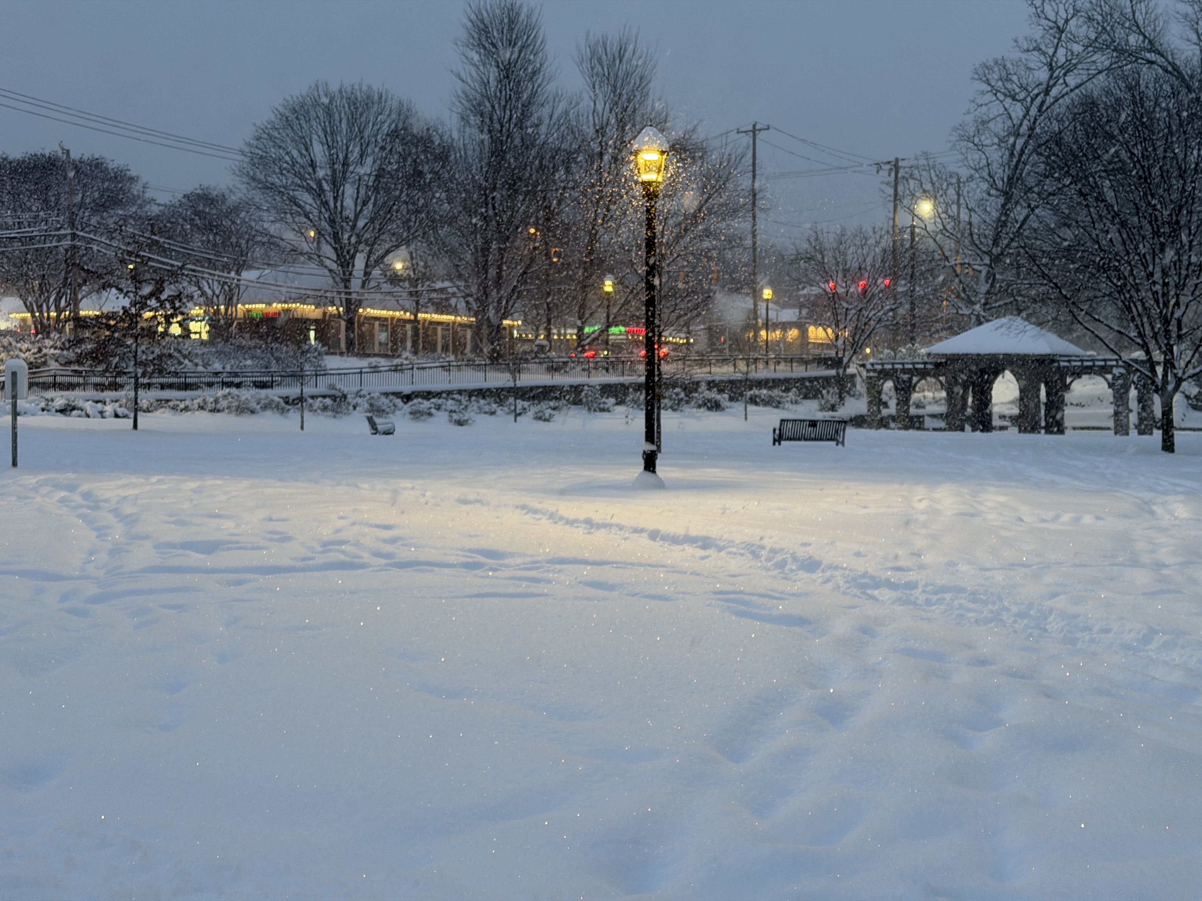 Snow-covered park with footprints, benches, a lit streetlamp, and a gazebo, with bare trees and a snowy street with lights in the background at dusk.