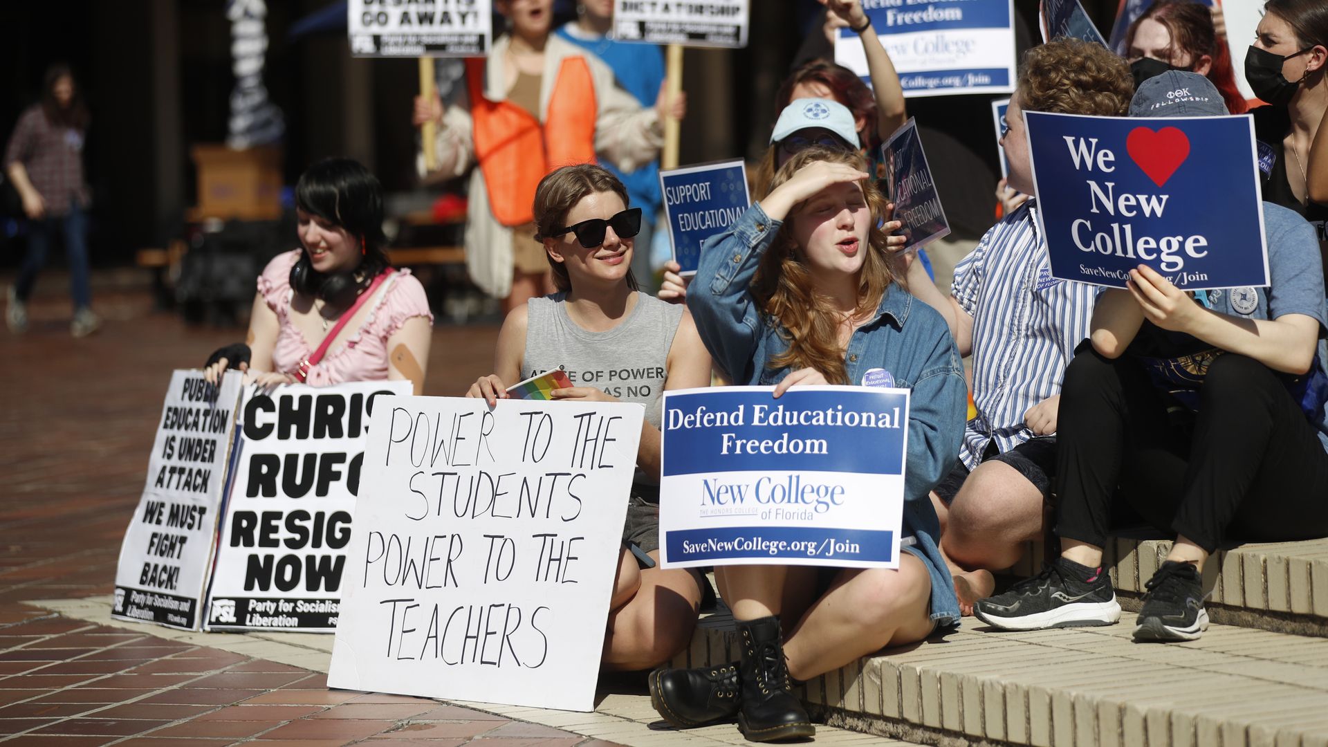 Supporters of New College of Florida sit on a curb holding protest signs. Among the sign messages: "POWER TO THE STUDENTS POWER TO THE TEACHERS," "Defend Educational Freedom," "CHRIS RUFO RESIGN NOW" and "We <3 New College."