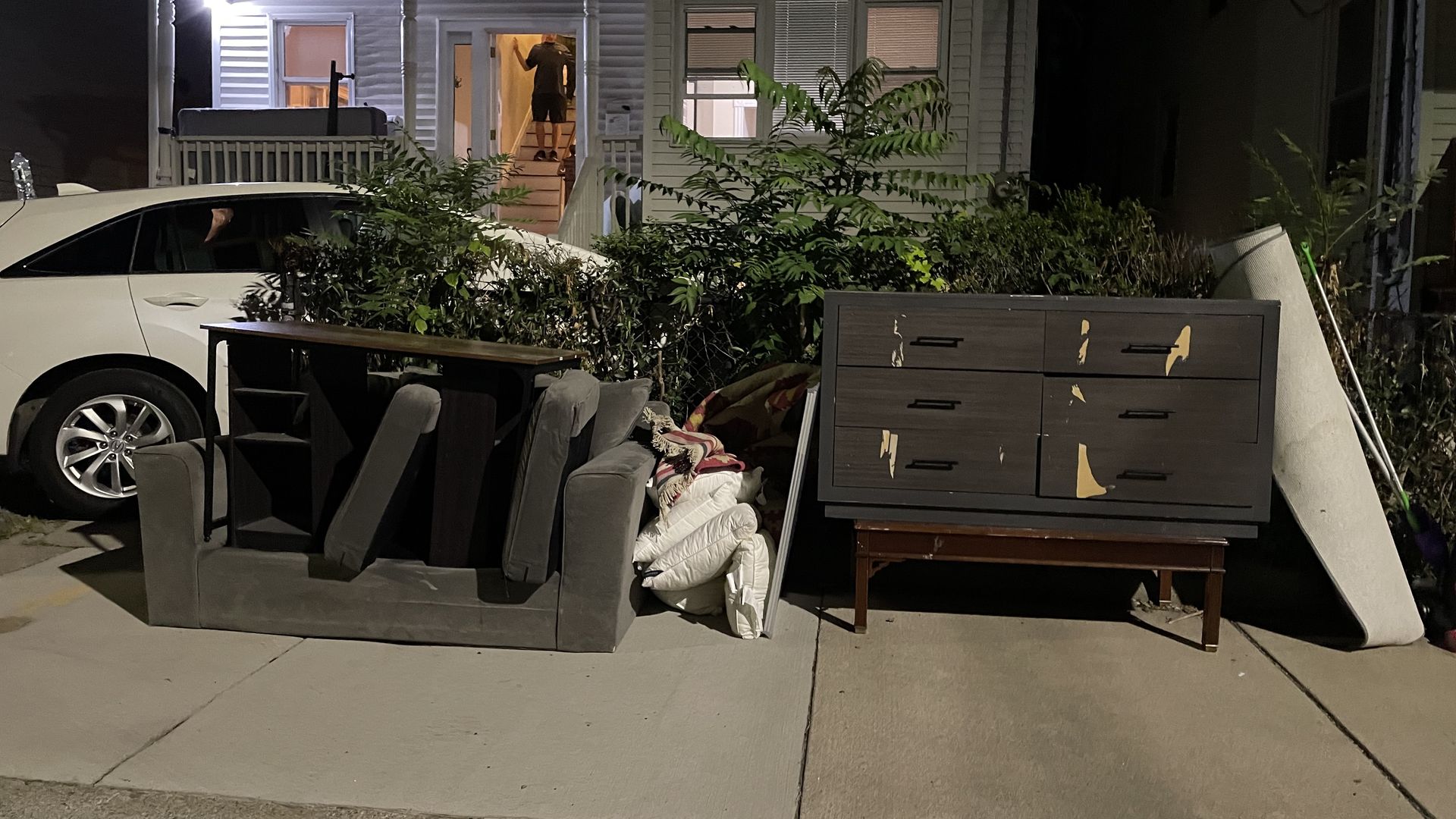 Night view of a house in Allston in late August, a white car parked on the left, and discarded furniture including an upside-down gray couch, drawers, mattress, and blankets on the driveway.