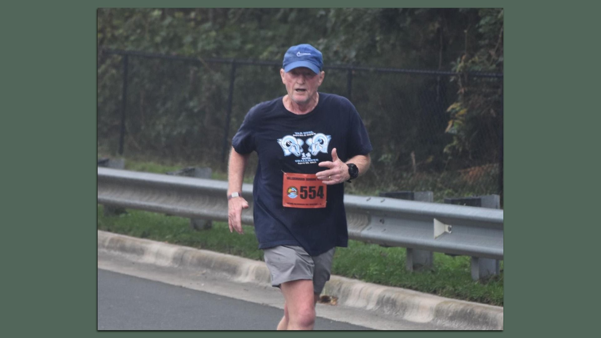 Older man running outdoors wearing a blue cap, navy T-shirt with ram logos and gray shorts, with race bib 554 on shirt near a roadside guardrail and trees in the background.