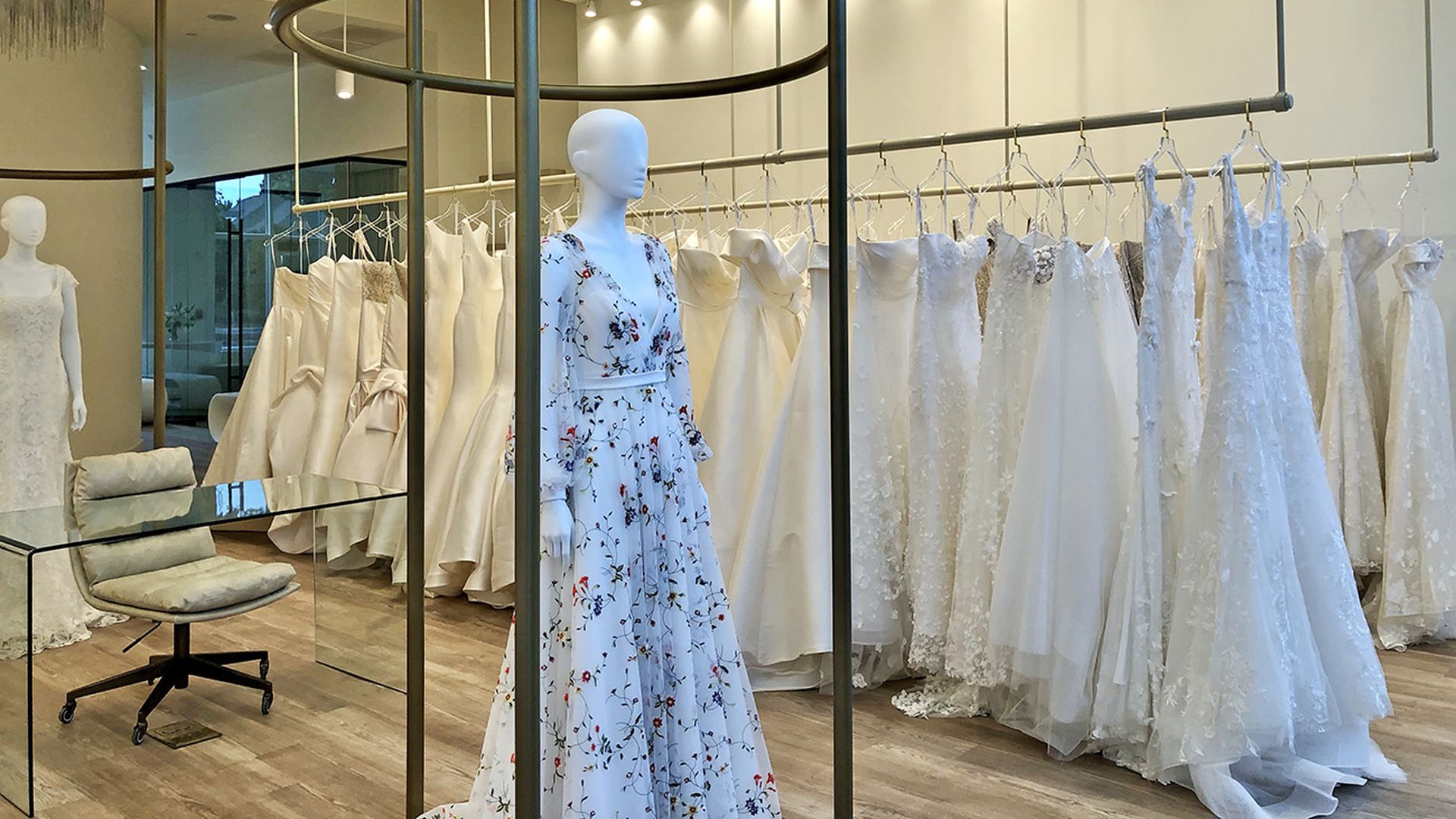 Elegant bridal boutique with white mannequins displaying wedding dresses; one mannequin wears a light blue floral gown; dresses in cream and white hang on gold racks under warm lighting.