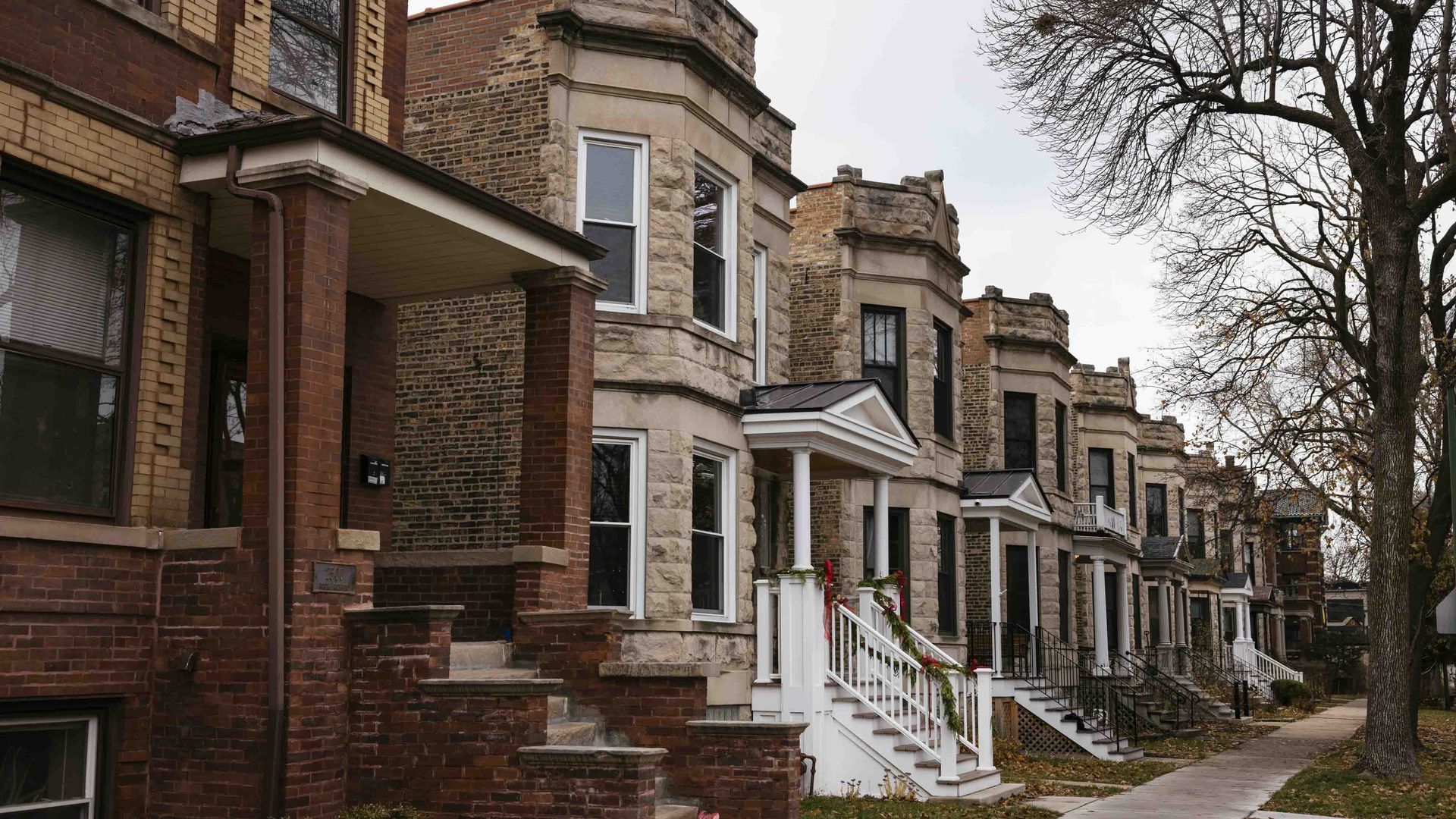 Row of vintage brick and stone houses with small front porches, some decorated with greenery, lining a sidewalk under a gray, overcast sky with leafless trees nearby.