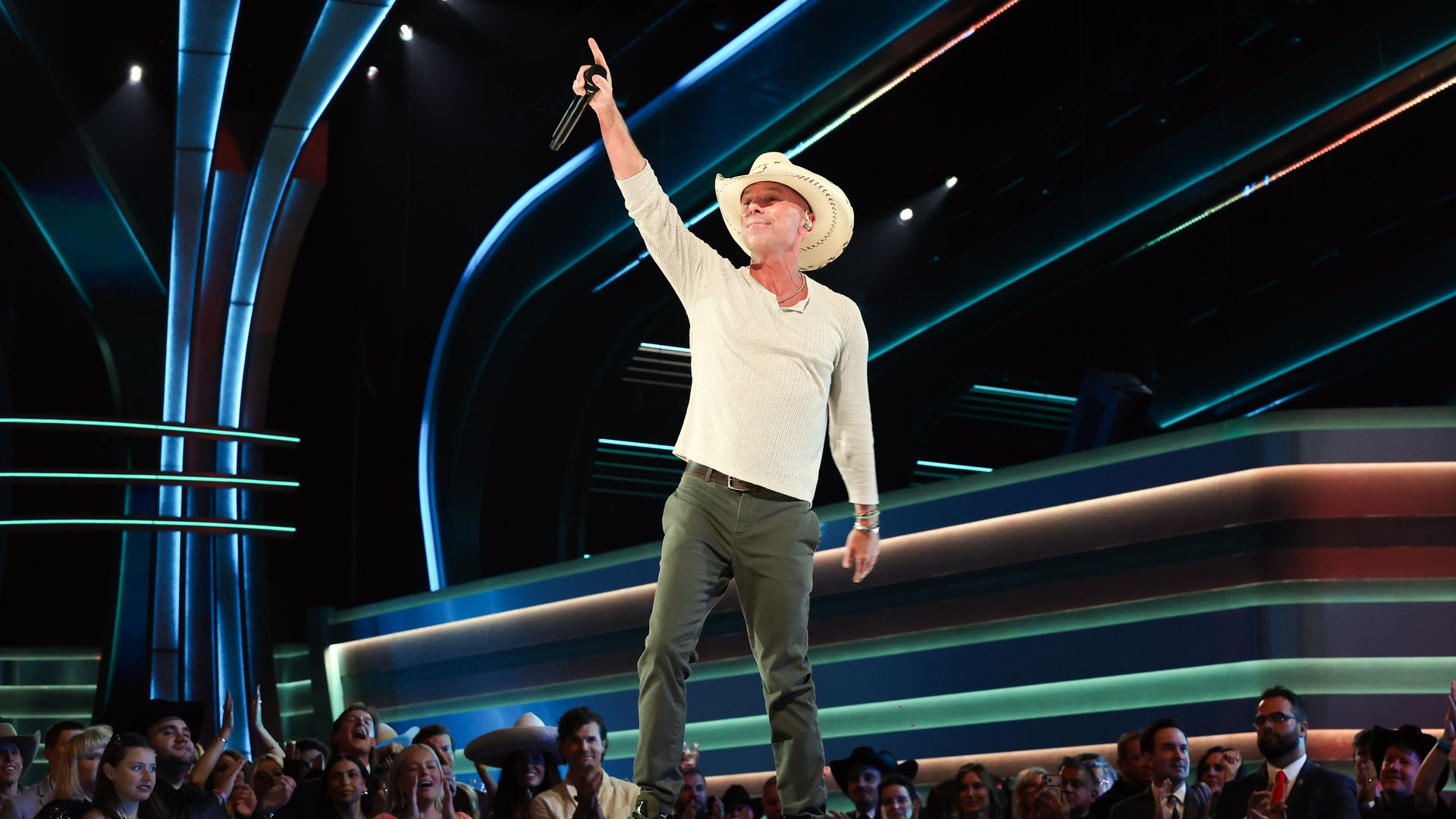 Kenny Chesney on stage with a cream long sleeved henley and a cowboy hat and green pants holding a mic in the air. 