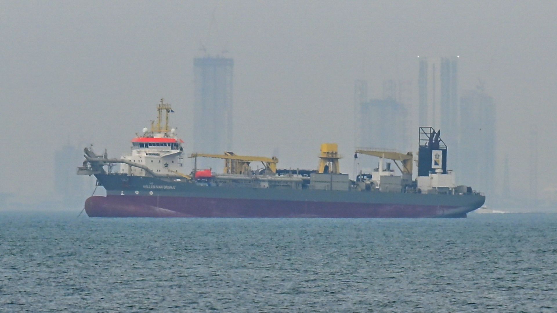 A large cargo ship with a green hull and red bottom, yellow deck cranes, sails on calm blue water; a hazy city skyline with tall buildings looms in the distance.
