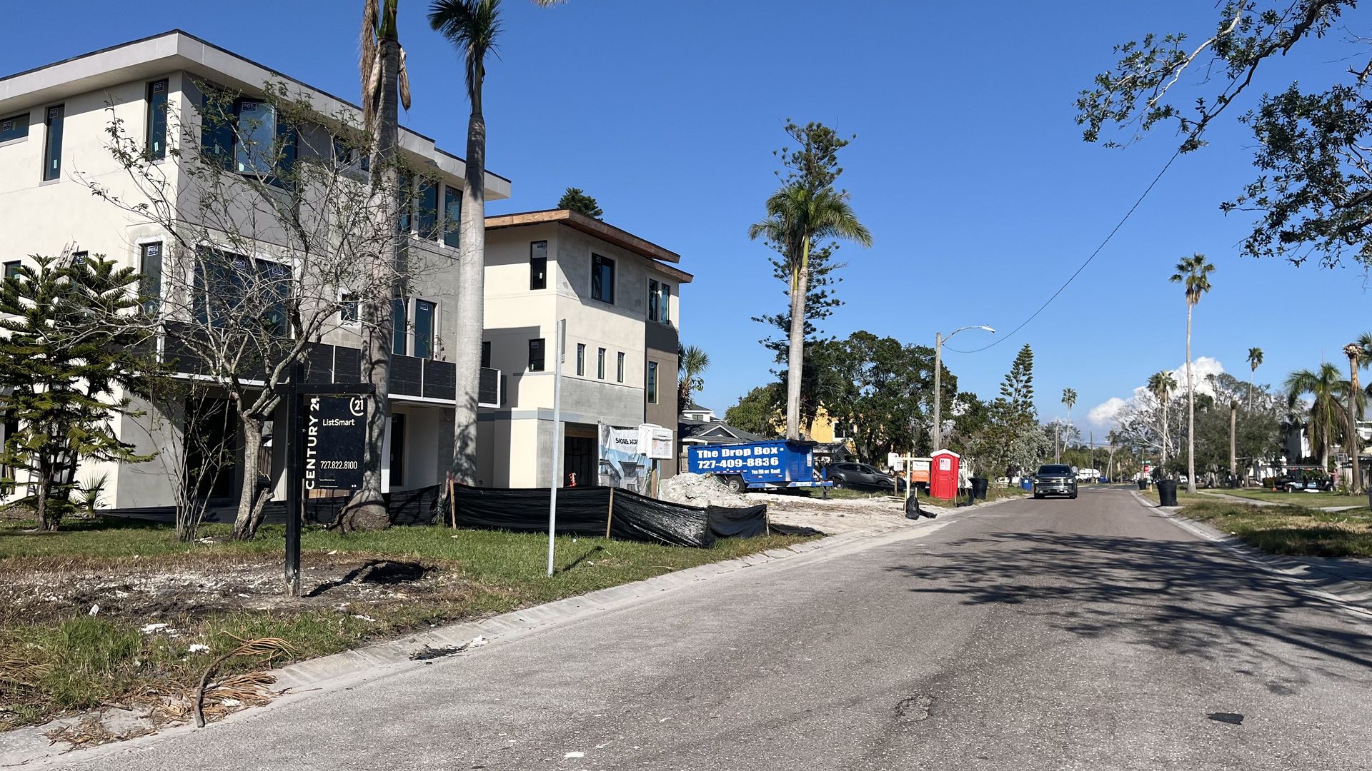 Sunny street with modern beige buildings under construction on the left, tall palm trees, and a clear blue sky. For-sale signs, a blue truck, and a red portable toilet sit by the curb.