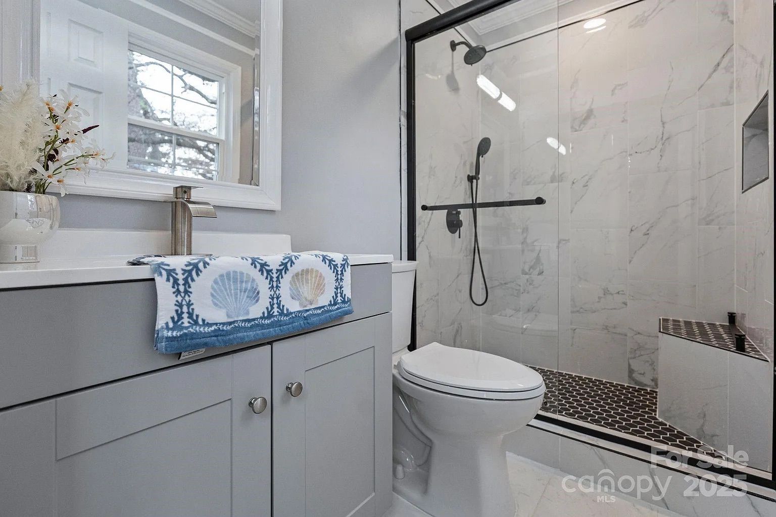 Modern bathroom with gray vanity, white countertop, decorative towel with blue seashells, white toilet, and glass-enclosed shower with dark hexagonal floor tiles and marble walls.
