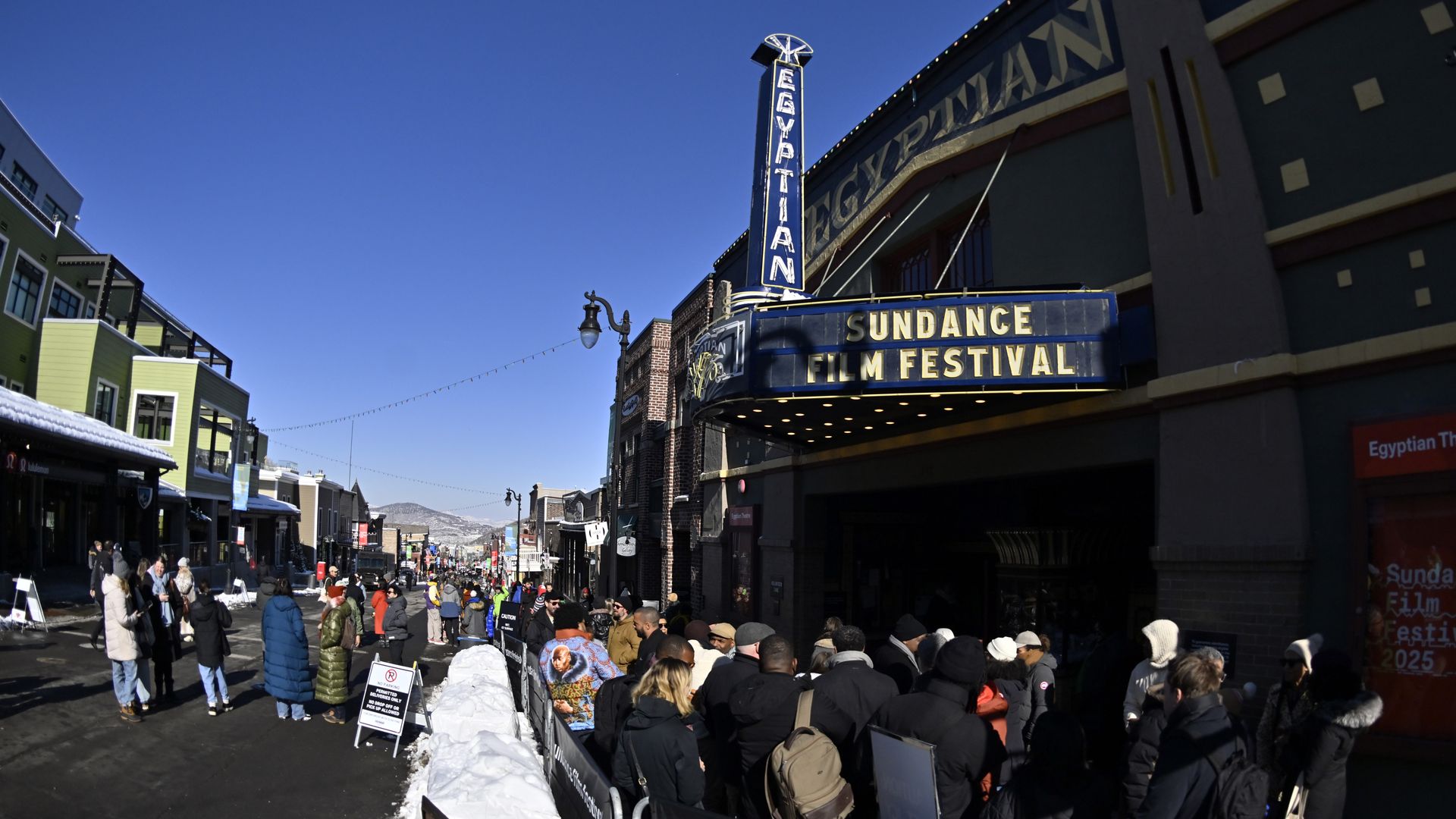 Crowd in winter coats lining up outside the Egyptian Theatre under a blue sky for the Sundance Film Festival, with snow on the ground and mountains in the background.