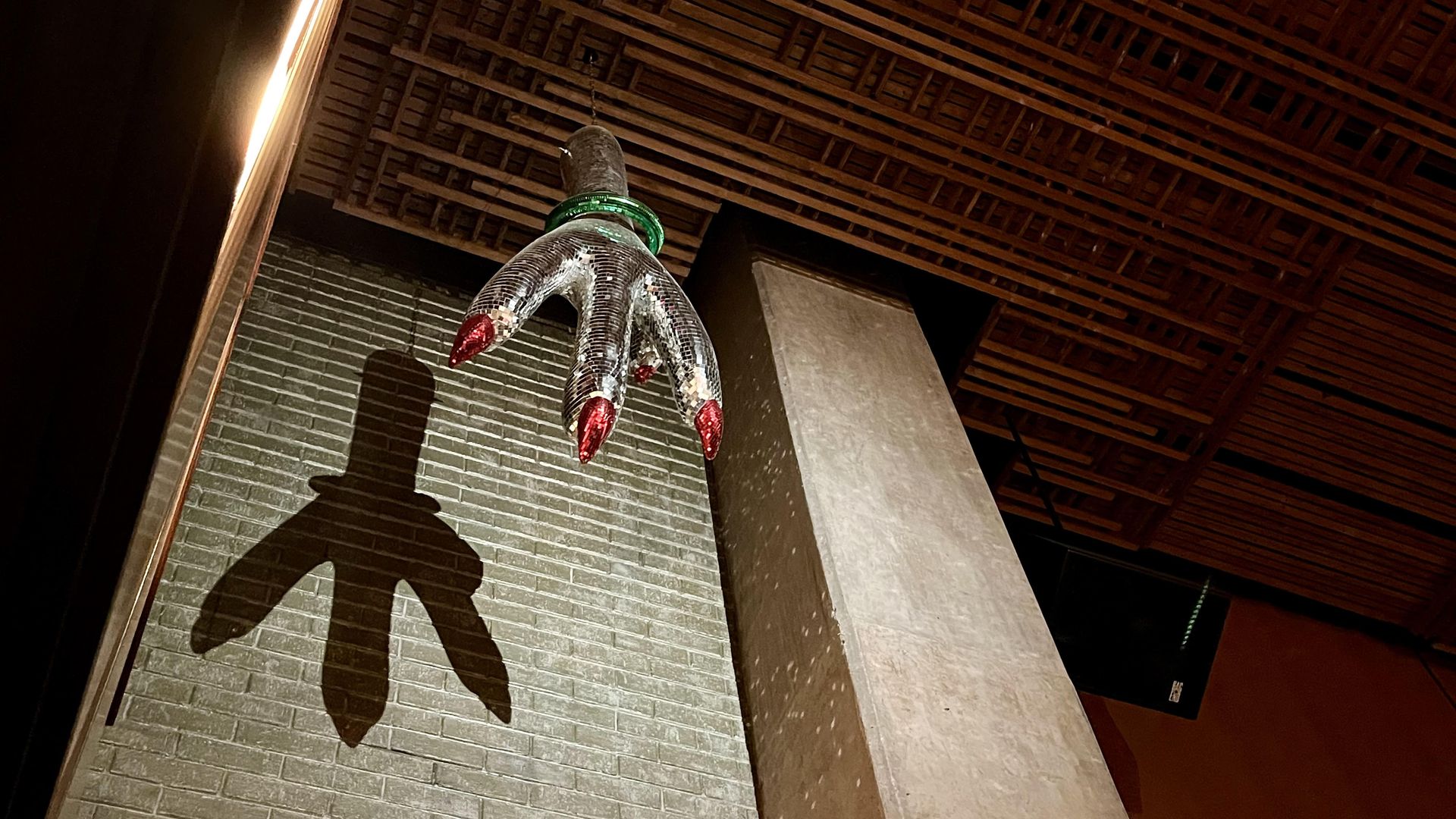 A ceiling disco ball installation of a large mirrored chicken foot with red claws and a green bracelet, casting a shadow on a brick wall, under a wooden lattice ceiling at Jue Let at Pearl.